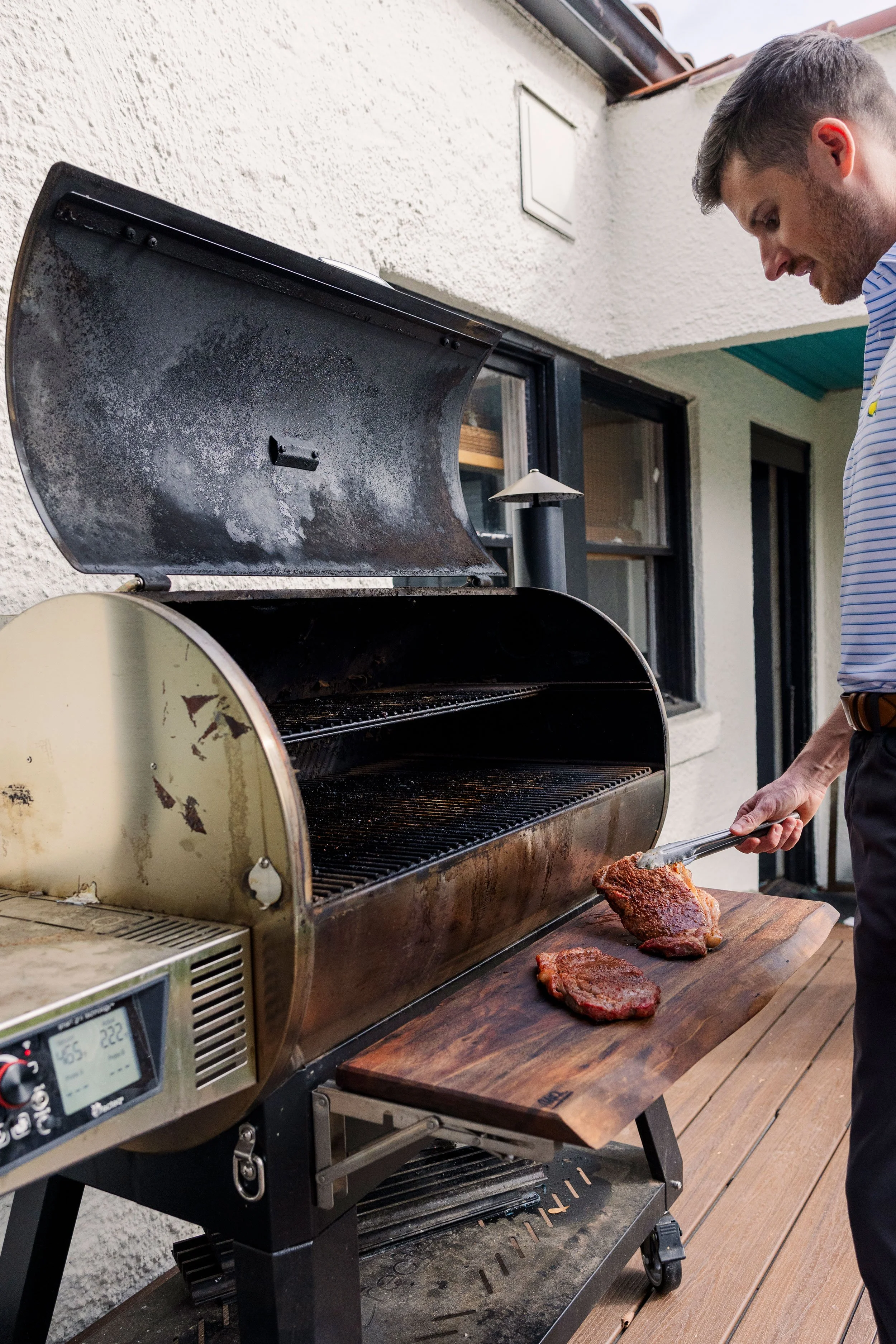 A man grilling meat on an outdoor barbecue grill on a wooden deck, with a white exterior wall and windows in the background.