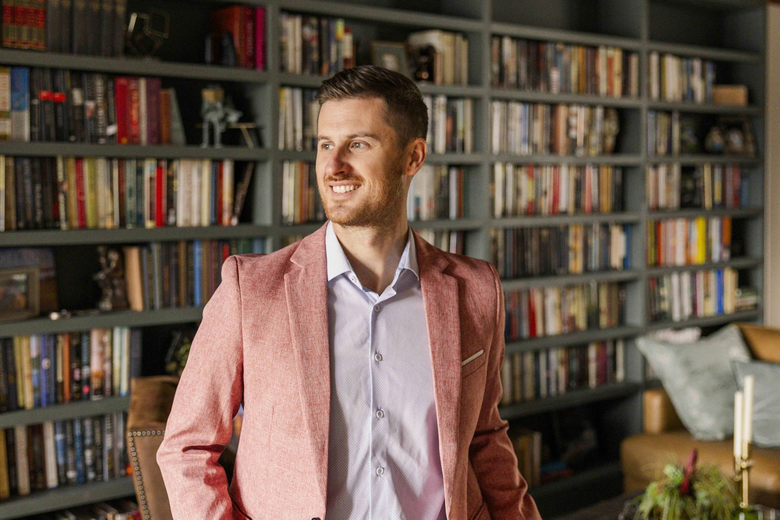 A young man with short brown hair, a beard, and wearing a pink blazer over a light blue collared shirt, standing and smiling while looking to the side in front of a large bookshelf filled with various books.