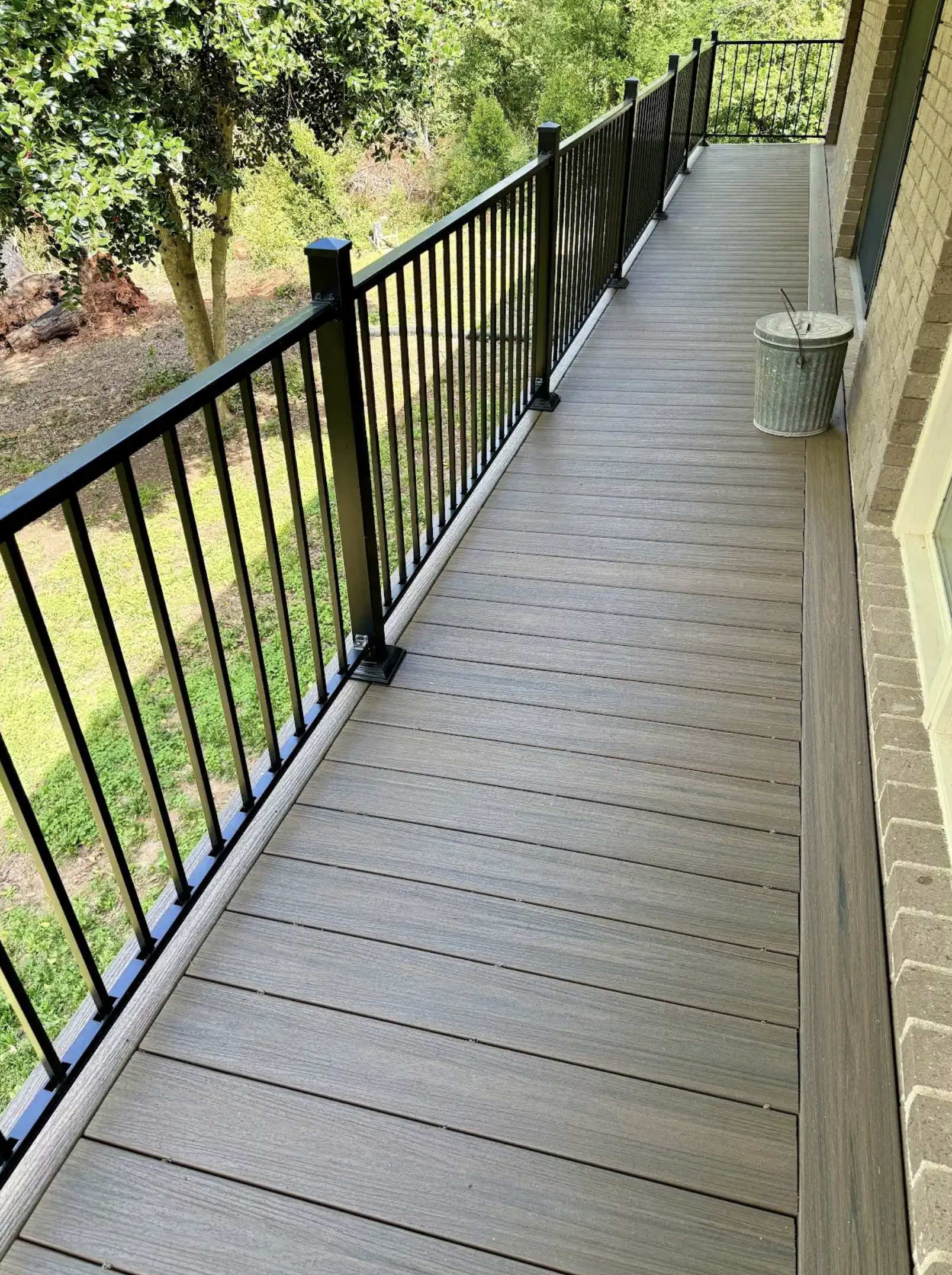 View of a balcony with brown wooden floorboards and a black metal railing, attached to a brick building. A metal trash can with a lid is placed on the balcony. There are trees and greenery visible beyond the railing.