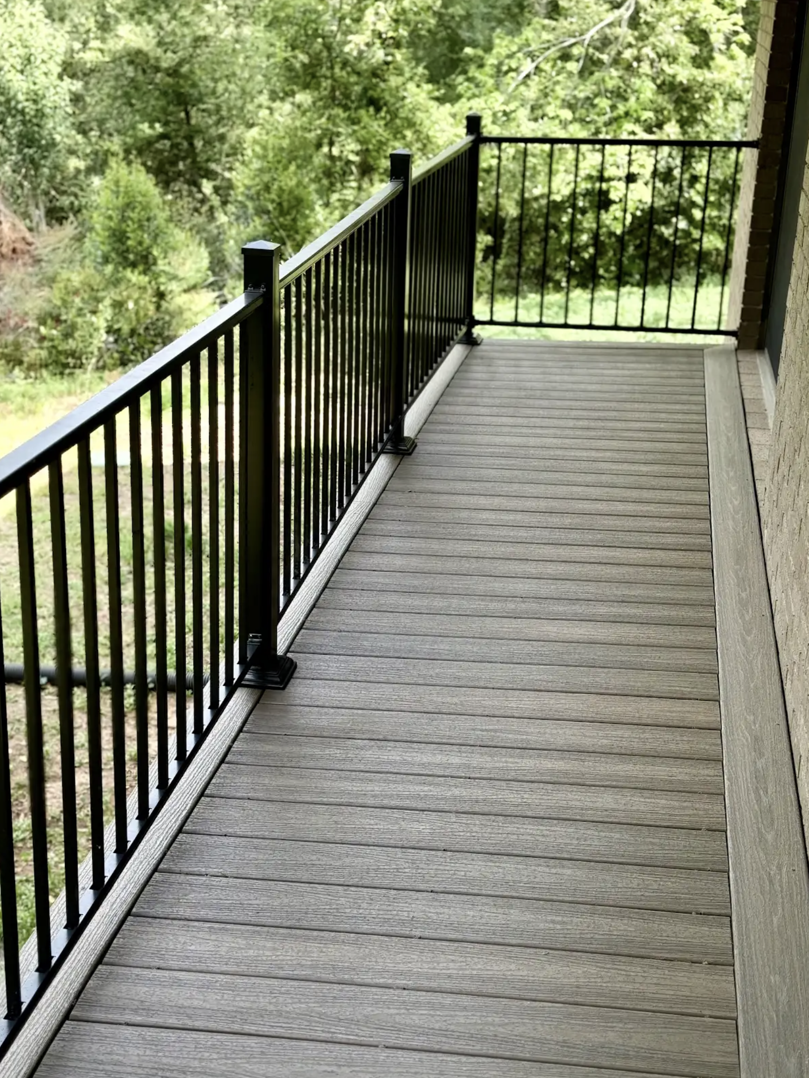 A balcony with wooden flooring and black metal railing overlooking a green wooded area.