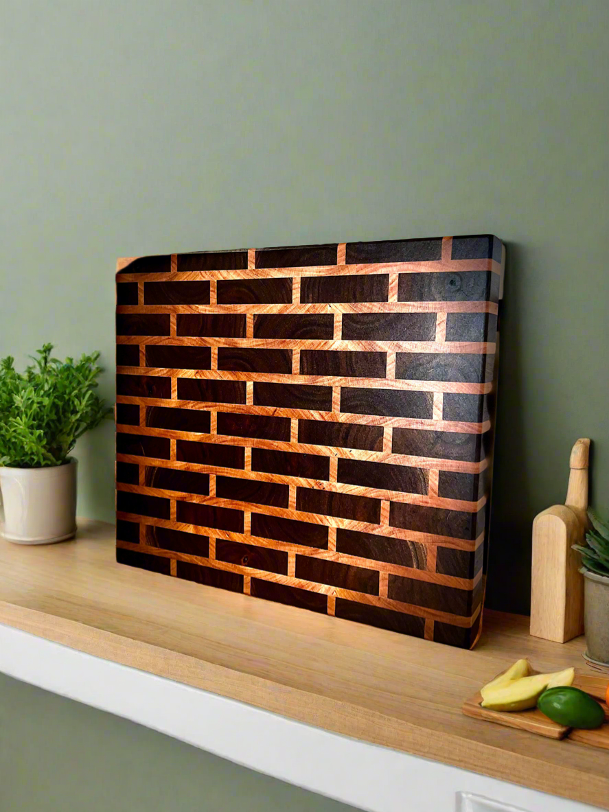 Wooden cutting board with a brick pattern design placed on a kitchen countertop, next to potted plants and a cutting board with bananas and a cucumber.