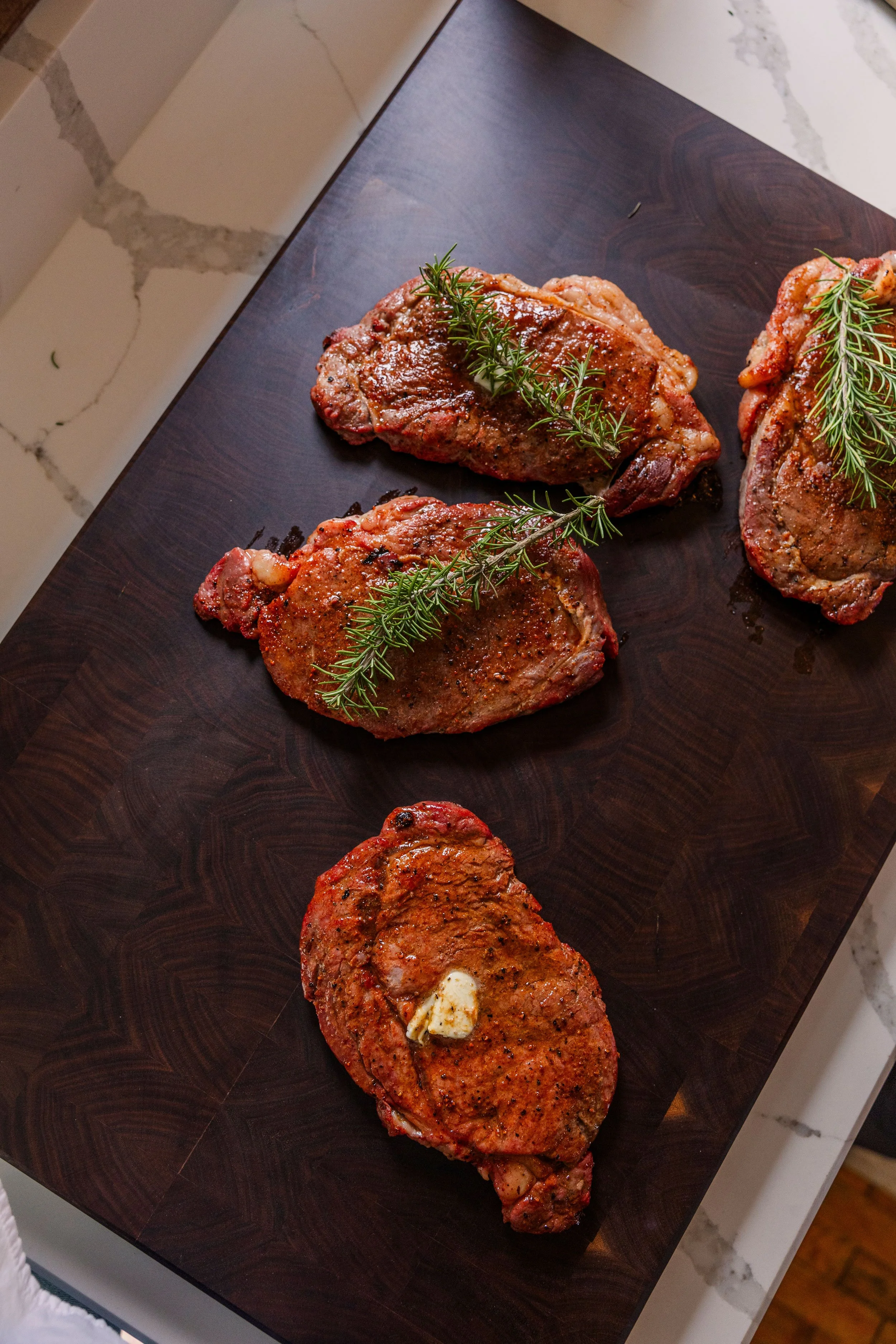 Three seasoned, raw steaks garnished with sprigs of fresh rosemary on a dark wooden cutting board.