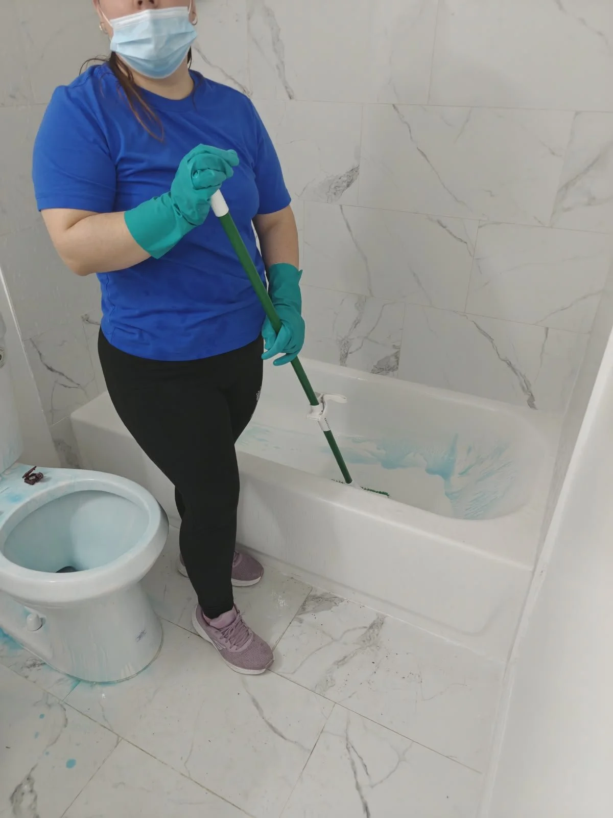 Persona limpiando la bañera con un trapeador en un baño, usando mascarilla, guantes y ropa deportiva.