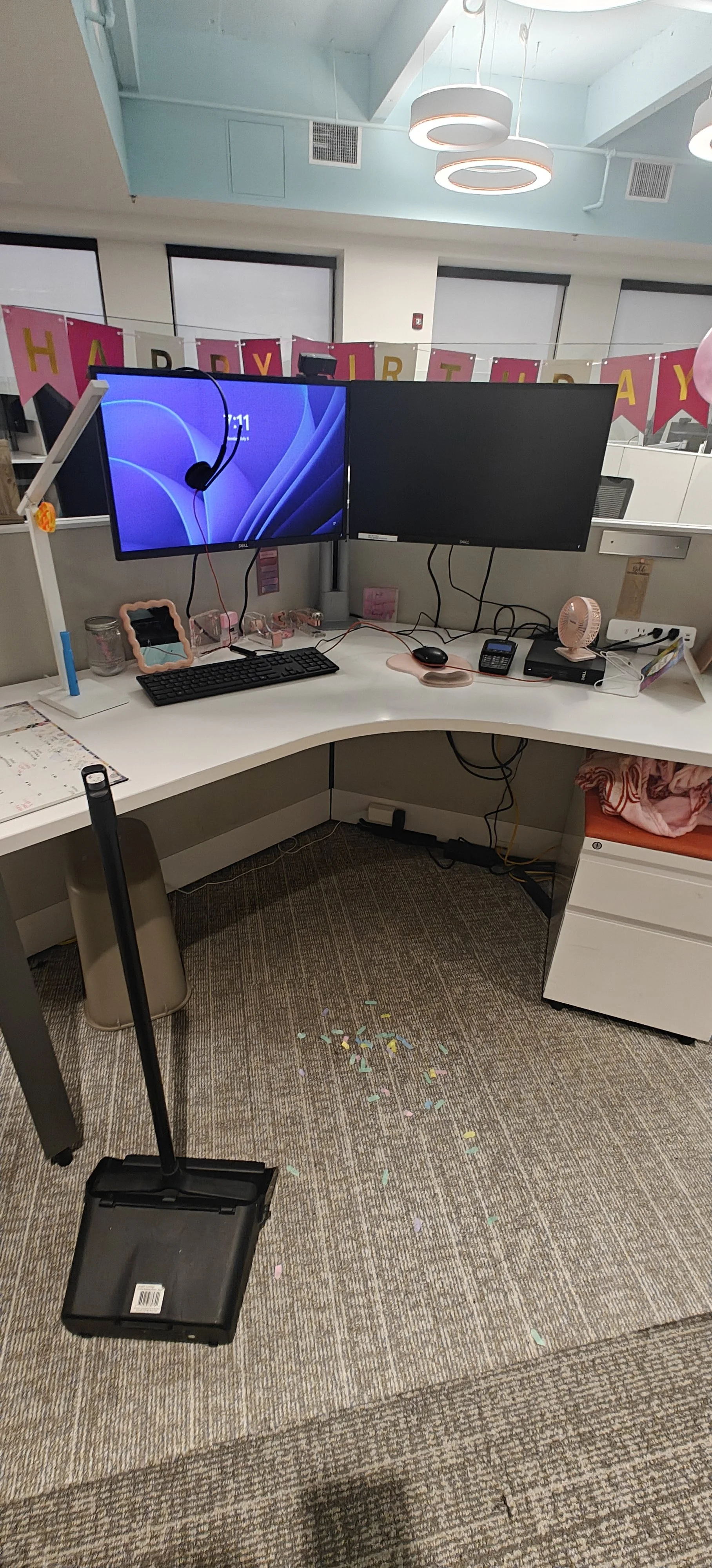 Una estación de trabajo de oficina con dos monitores, teclado, ratón, dos teléfonos, un ventilador pequeño, y decoraciones decorativas rosadas y una lona de cumpleaños en el fondo. Hay confeti en el suelo, y una escoba y un balde en primer plano.