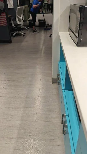 Oficina con personas limpiando y muebles de oficina, incluyendo una estantería azul y una televisión en la esquina.