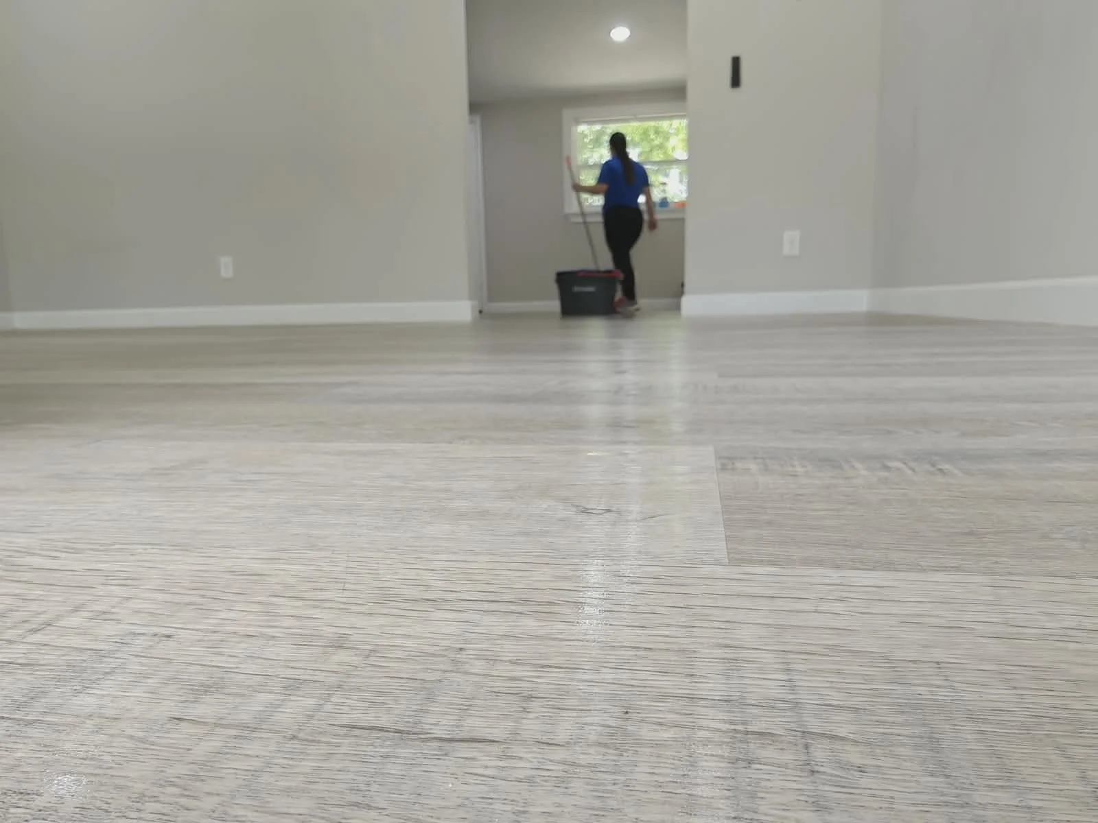 Persona limpiando un suelo de madera clara con trapeador, vista desde nivel del suelo en una casa moderna con paredes grises y ventanas grandes.