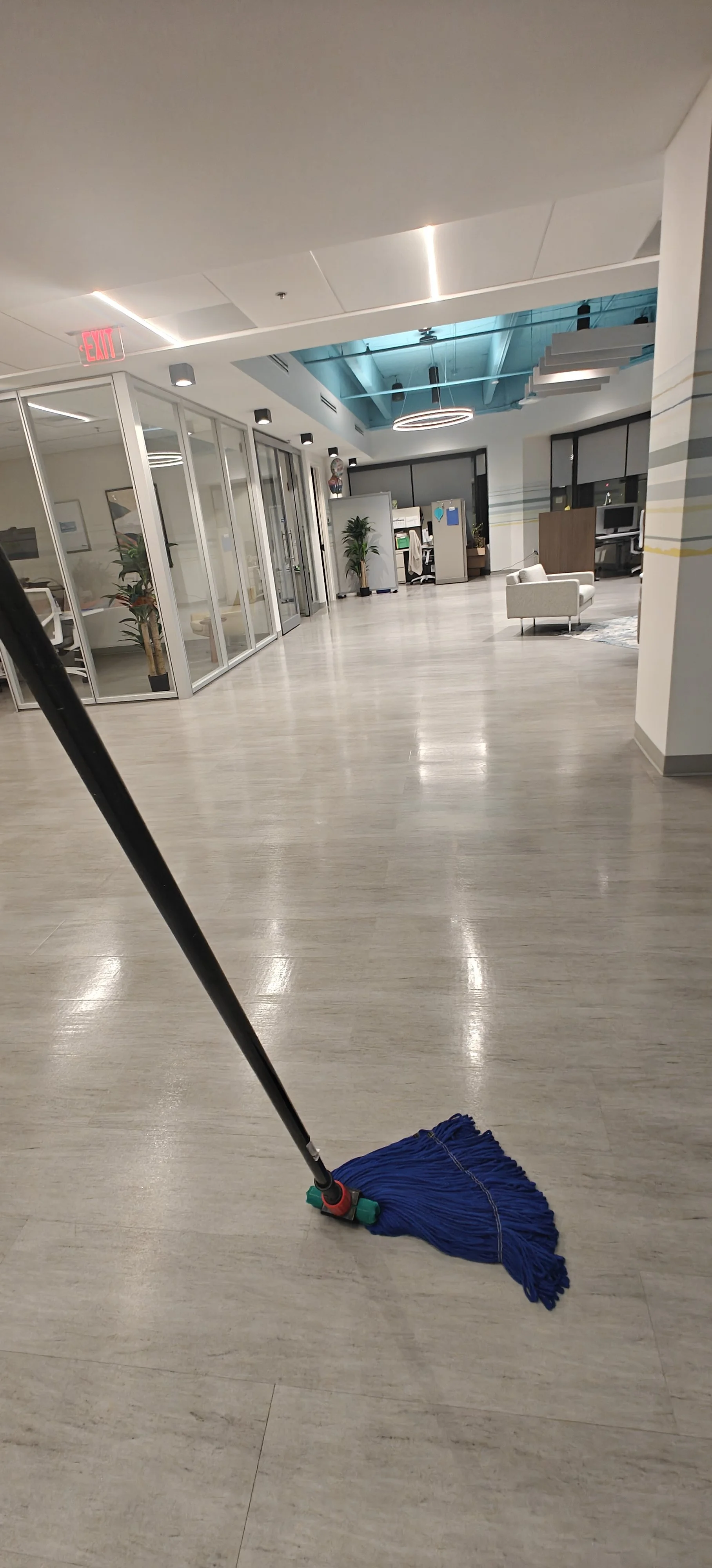 Sala de oficina vacía con piso de madera clara y un área de descanso con sillones blancos en el fondo. En primer plano, una mopa azul en el suelo, con un mango negro y una base roja y verde.