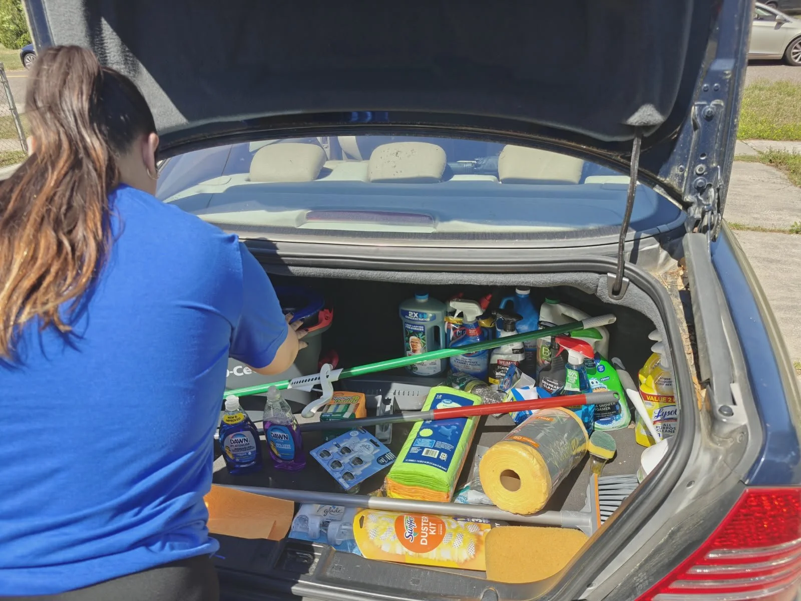Una mujer con camiseta azul organiza productos de limpieza en el maletero de un coche, que contiene detergentes, toallas de papel y otros artículos de limpieza.