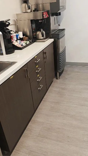 Mostrador de kitchenette con cafetera, tetera y dispensador de agua en una oficina o área de descanso.