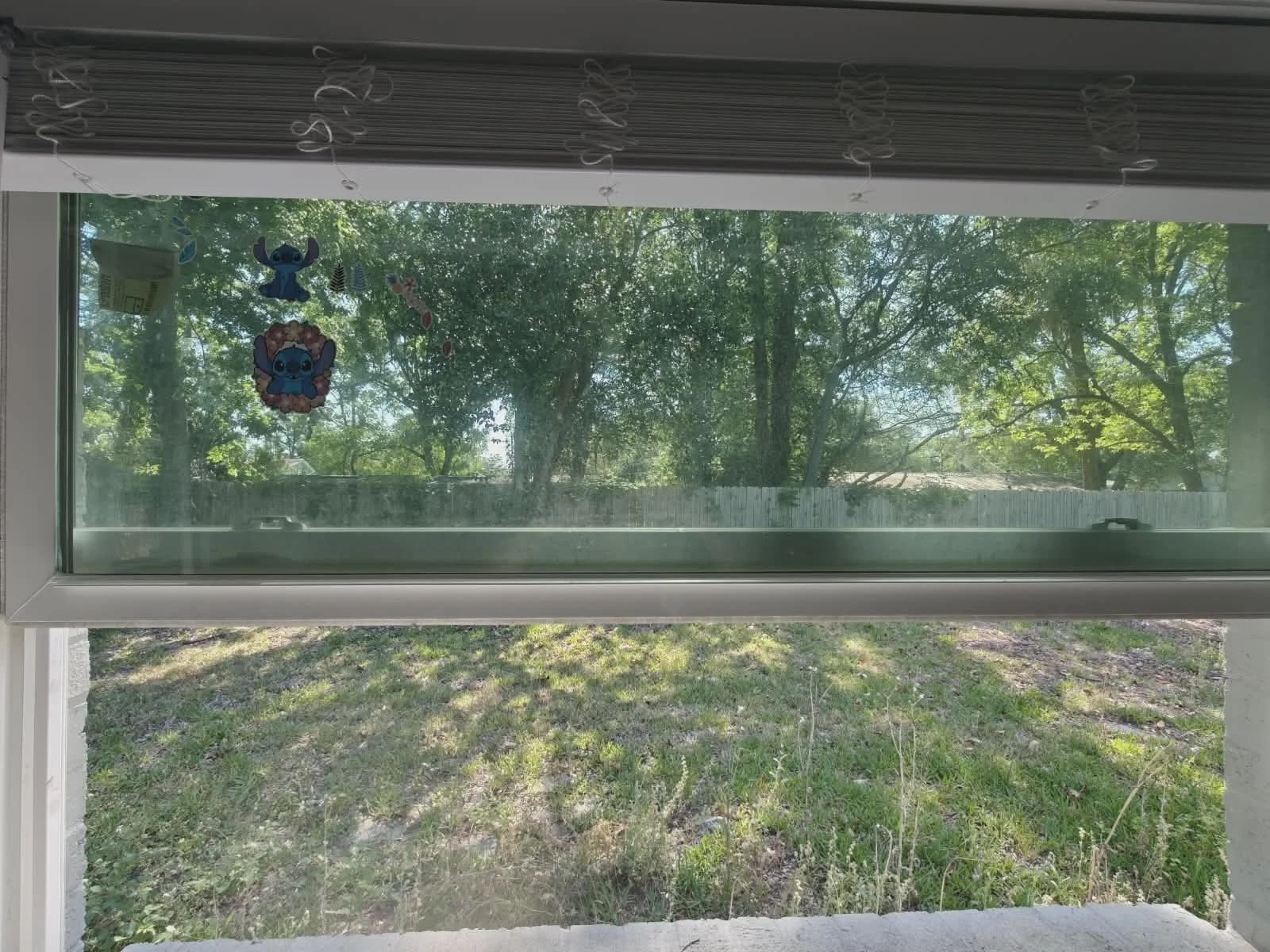 Ventana con cortinas blancas y decoraciones de personajes de Stitch en la parte de afuera, vista hacia un jardín con árboles y césped.