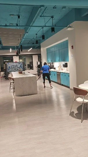Área de cafetería o comedor con mobiliario moderno, paredes blancas y cocinas azules, algunas personas sirviéndose comida o bebidas.