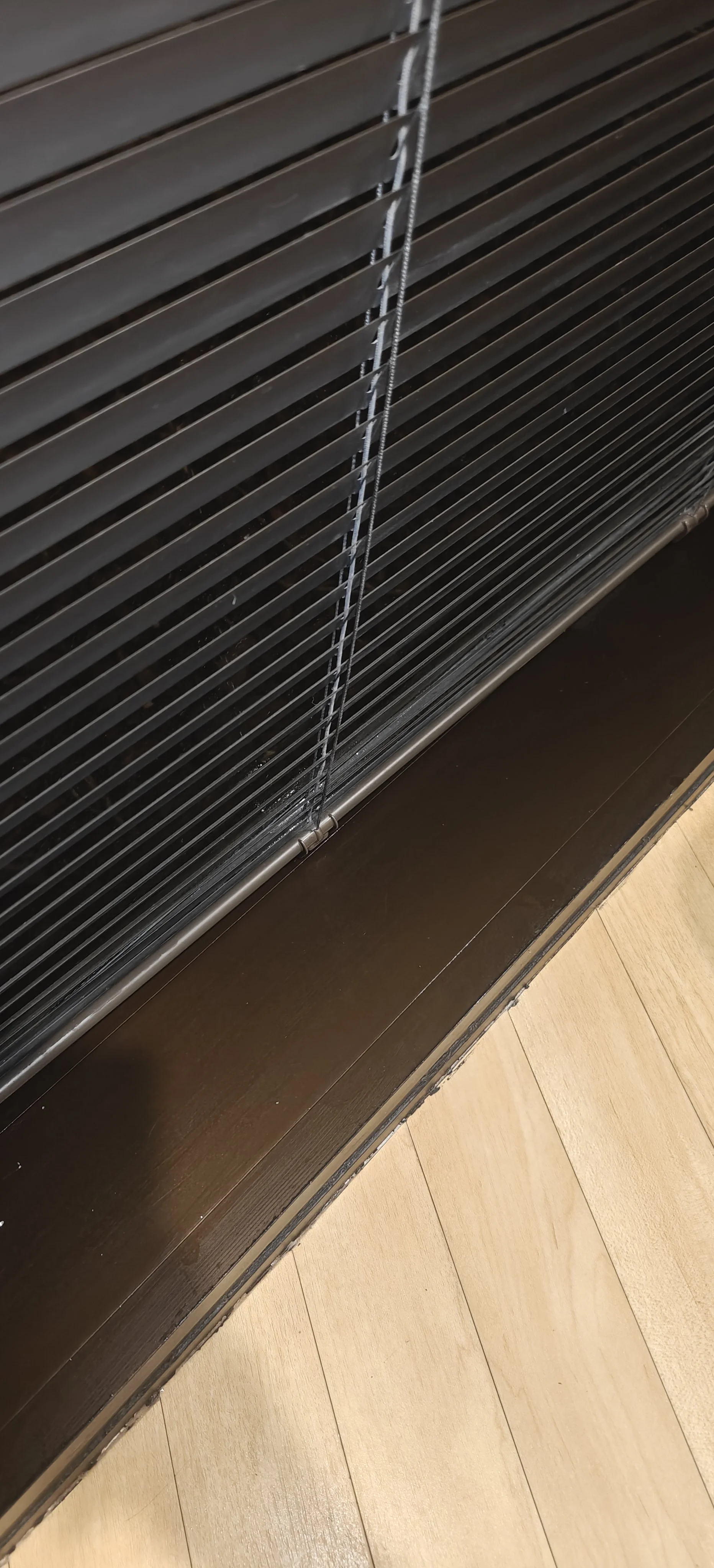 Reja metálica de cortina enrollable con cuerda de control, instalada en una ventana o puerta, sobre un marco de madera y piso de madera claro.