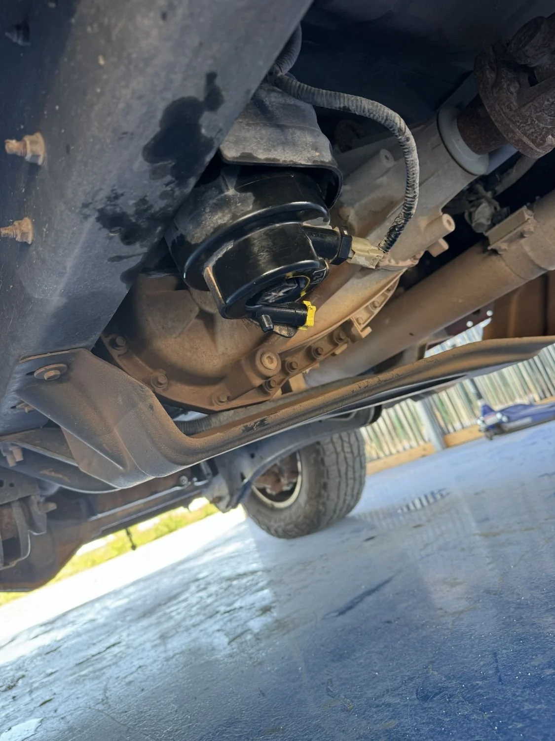 Underneath view of a vehicle showing a black electric fuel pump attached to the underside of the vehicle's engine or transmission area, with various hoses and connectors visible.