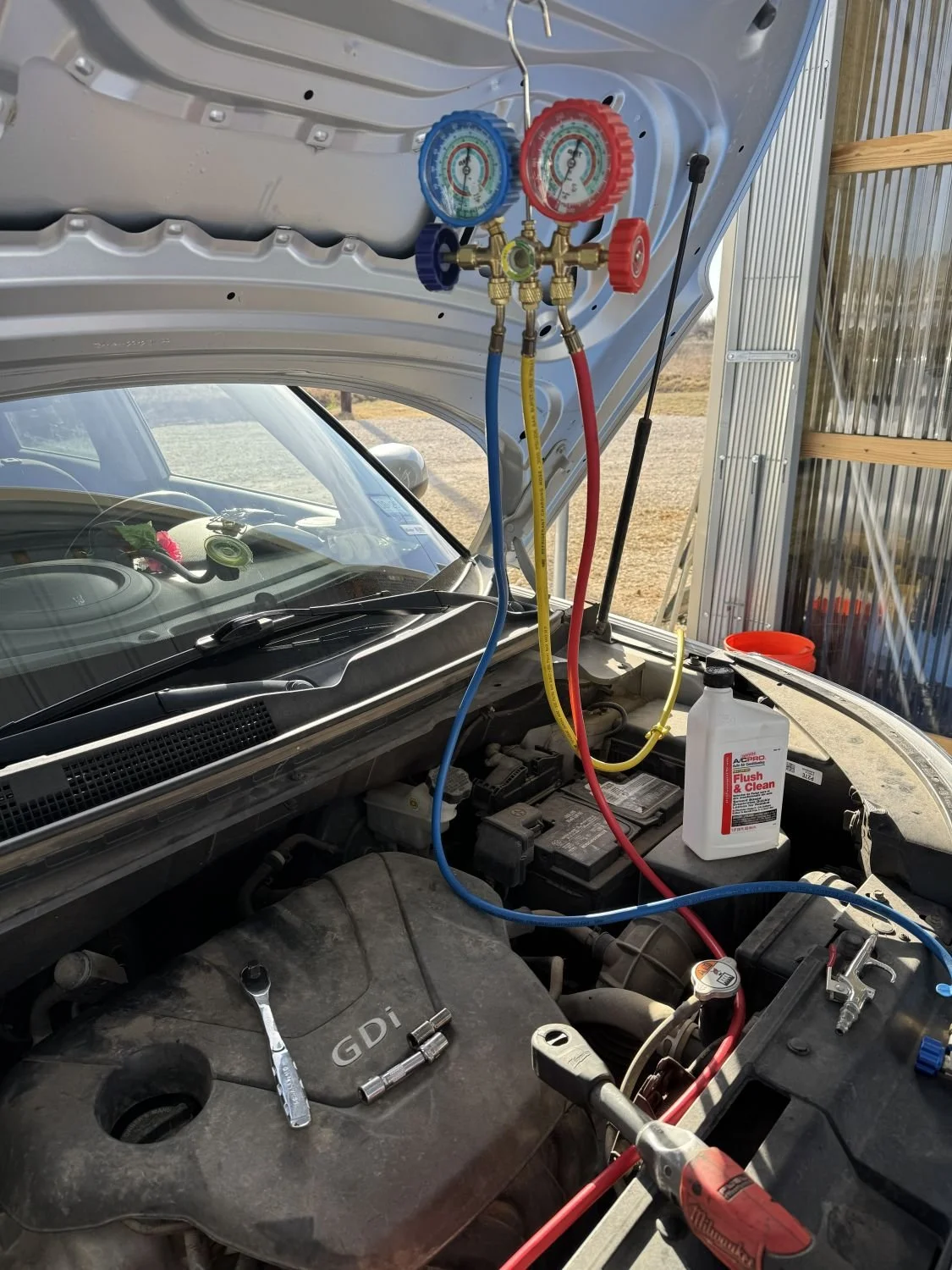 Vehicle hood open showing HVAC manifold gauge set connected to the air conditioning system, with hoses attached to the refrigerant ports, and tools on the engine.
