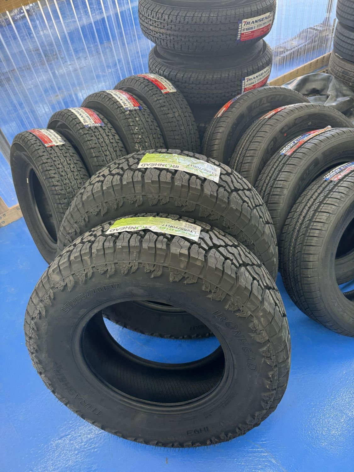 Stack of new tires arranged on a blue floor at a tire store, with some tires wrapped in labels and others stacked in the background.