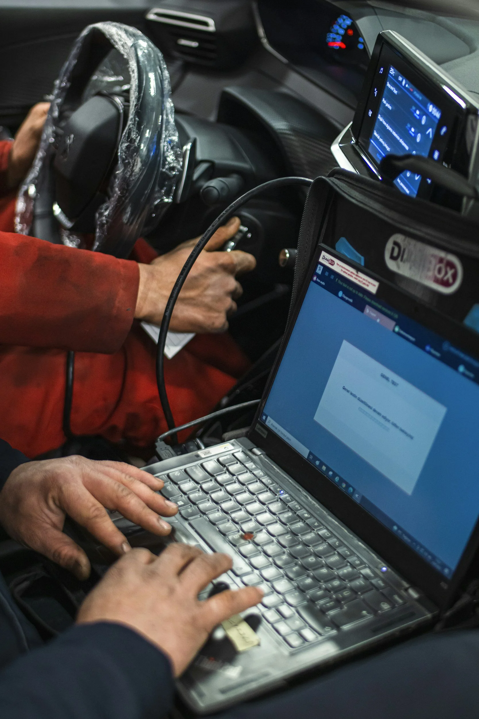 Inside a vehicle, a person wearing a red jacket types on a computer keyboard while another person holds a wrapped steering wheel. There are two screens displaying data; the one in the vehicle appears to be a tablet with a blue interface, and the other on a laptop shows a web page.