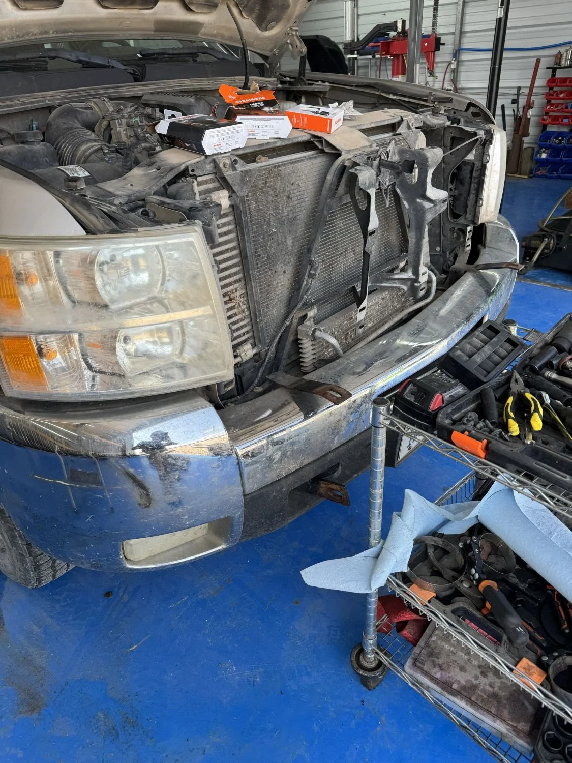 Front of a car with the grille and radiator removed, exposing the engine compartment, in a garage workshop. Tools and parts are on a nearby shelf.