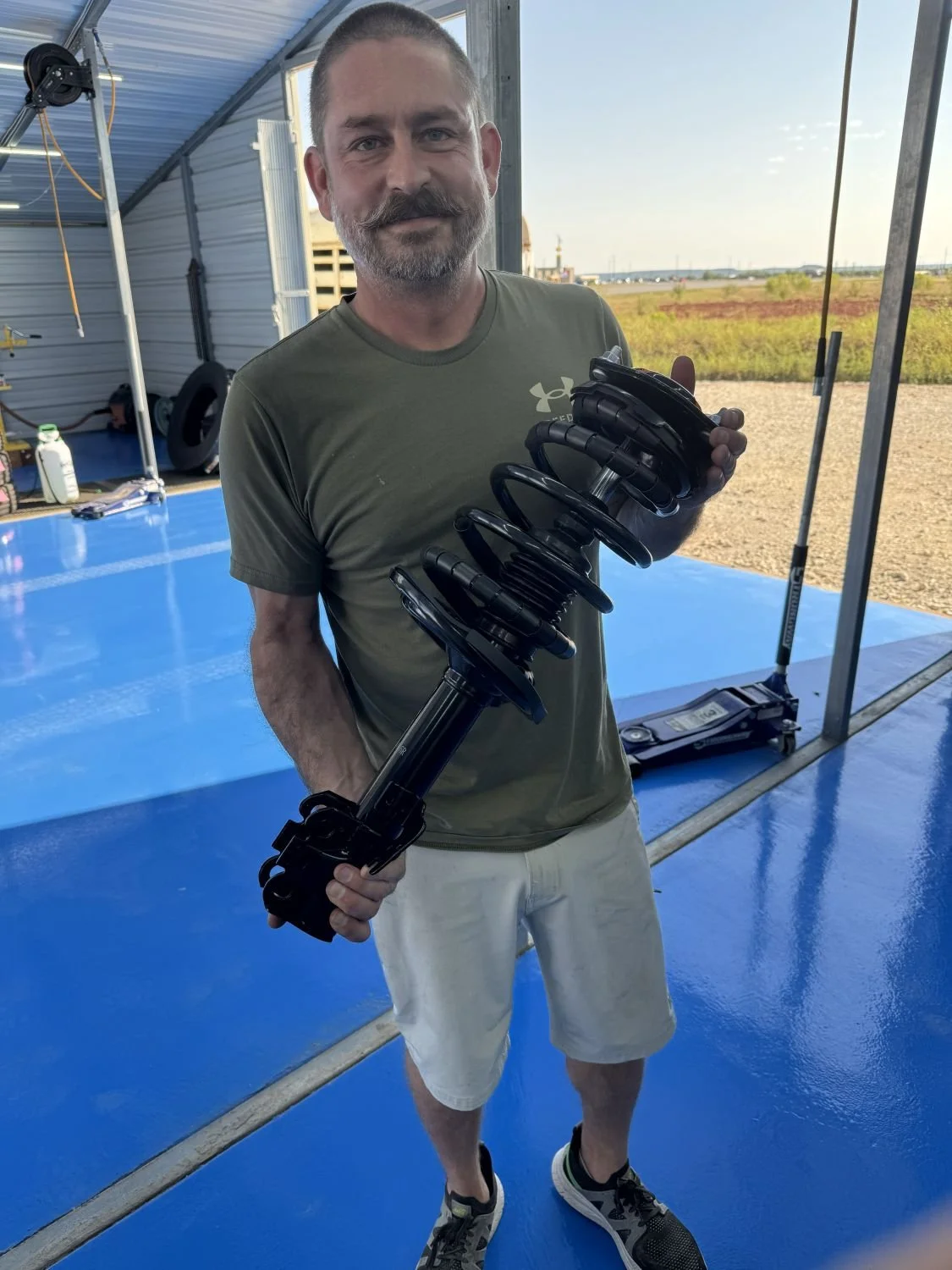 Man standing indoors holding a large black coil suspension part, wearing a green Under Armour t-shirt, light shorts, and sneakers, with a blue floor and outdoor landscape visible through the open garage door.