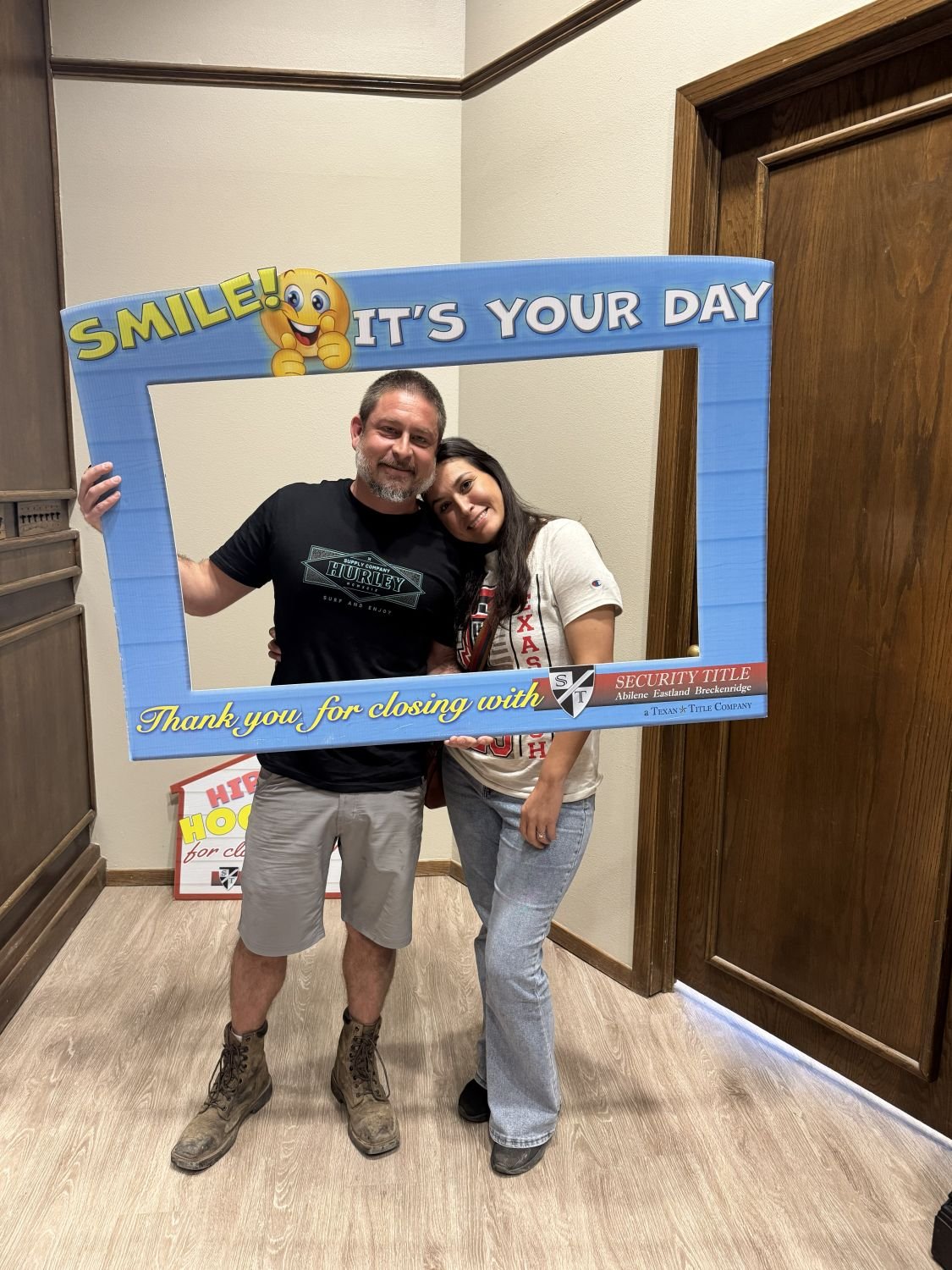 A man and woman smiling and hugging inside a room, holding a blue photo frame with the words 'Smile! It's Your Day. Thank you for closing with' and a security title company's logo.