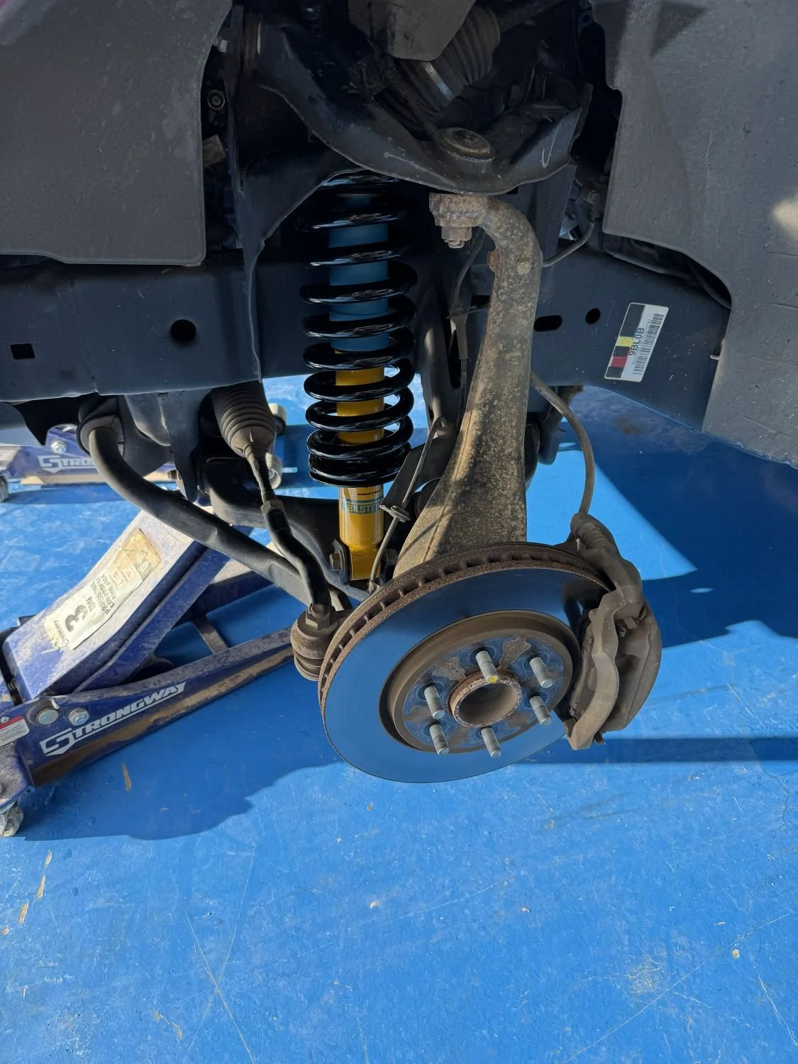 Auto repair shop with a vehicle lifted on a jack, showing the suspension system, brake rotor, and brake caliper assembly, with the wheel removed.