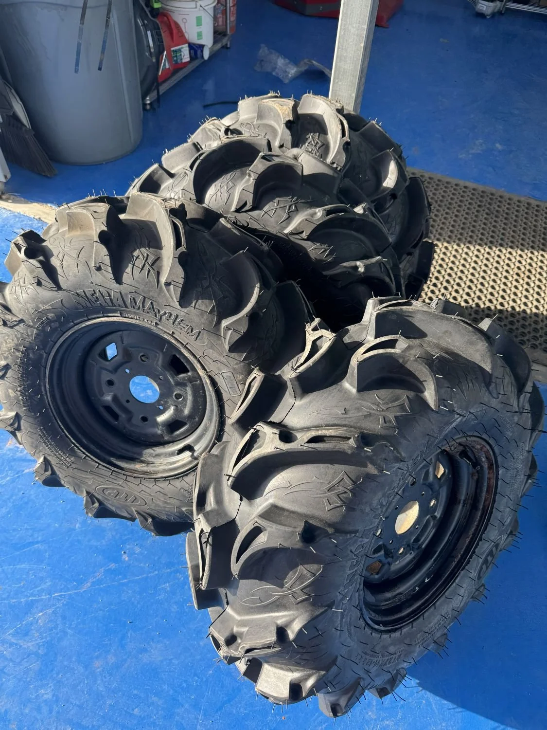 Group of four large, rugged off-road tires stacked together on a blue floor in a workshop or garage setting.