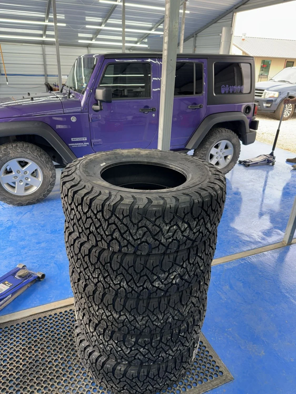A purple Jeep parked inside a garage, with three stacked large off-road tires and a tire jack on the blue floor nearby.