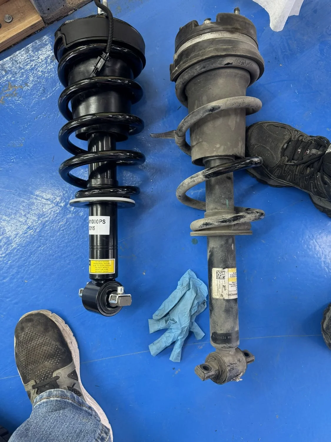 Comparison of a new black coilover shock absorber and an old dirty shock absorber on a blue floor, with a sneaker and a pair of blue gloves nearby.
