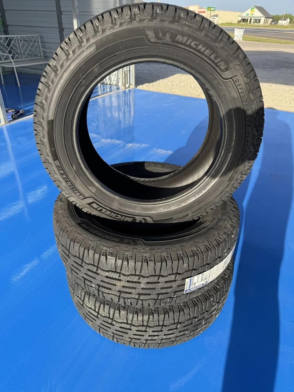 Two stacked black Michelin Defender T+X tires on a blue surface outdoors, with buildings and a road in the background.