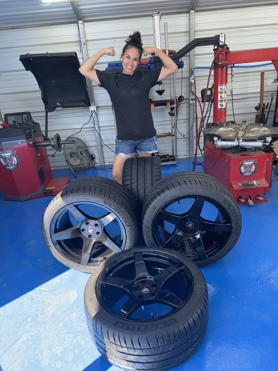 A woman in a black t-shirt and denim shorts flexing her arms in a garage, with four car tires and various automotive equipment and tools around her.