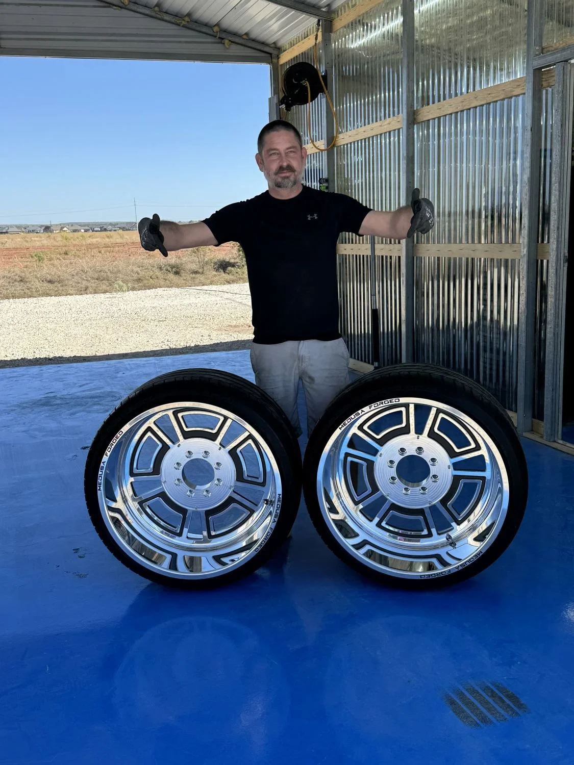 A man stands inside a garage with corrugated metal walls, smiling with his arms outstretched. In front of him are two silver alloy wheels with tires, labeled "MOUA FORGED." Outside the garage, there is a clear sky and open land.