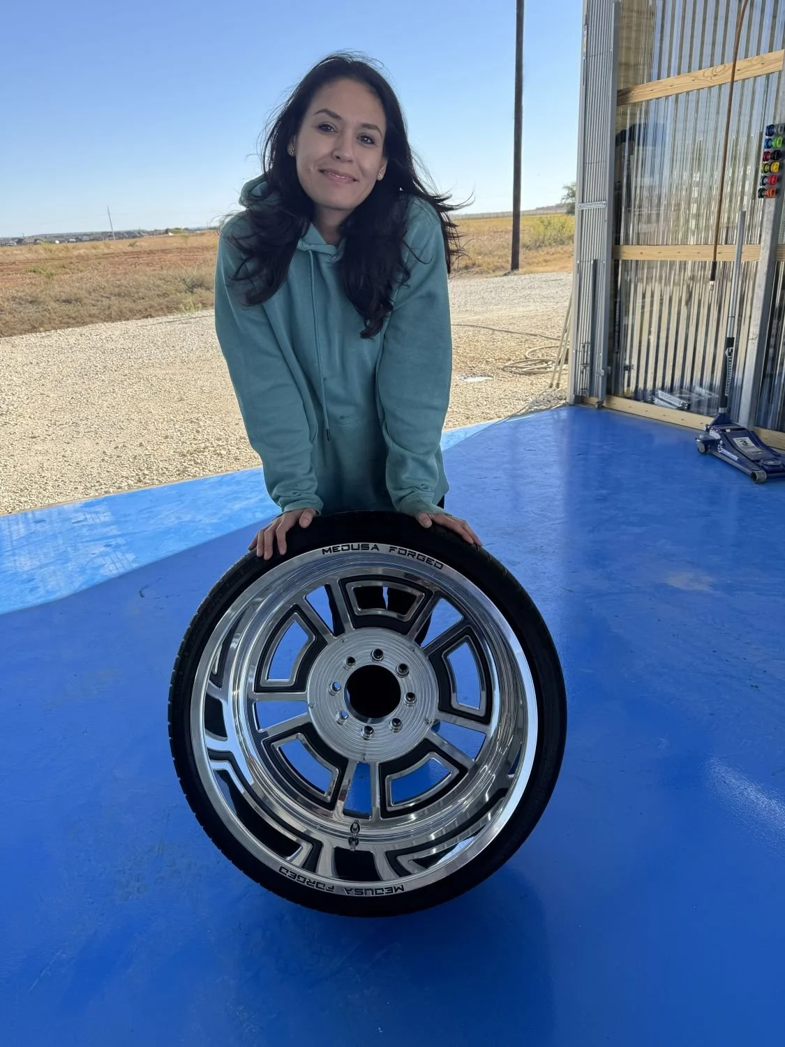 Woman in a teal hoodie leaning on a large car wheel with a blue floor and open garage or workshop setting outdoors.