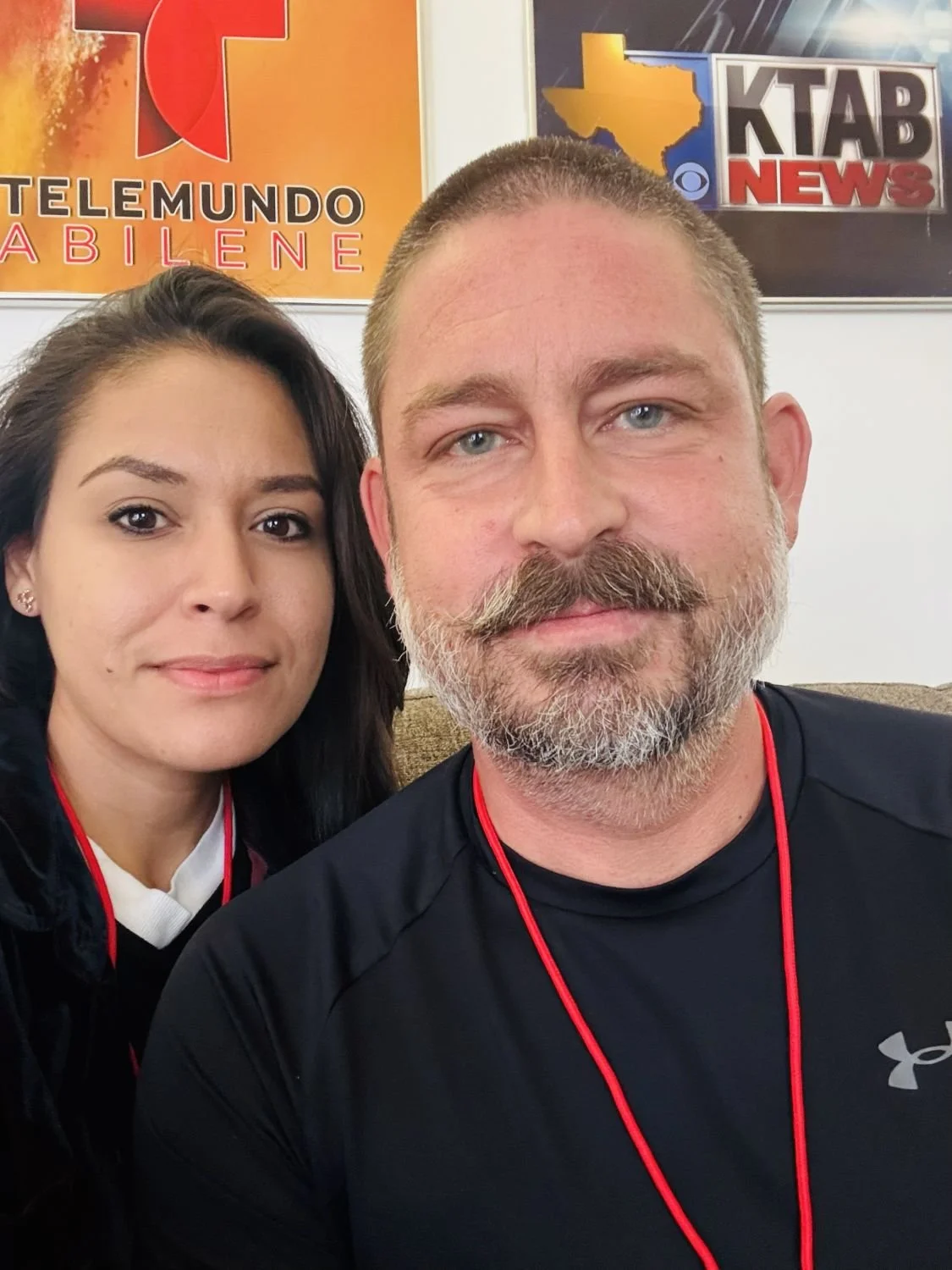 Close-up of a man with a beard and mustache, and a woman with dark hair, sitting together, with television news posters in the background.