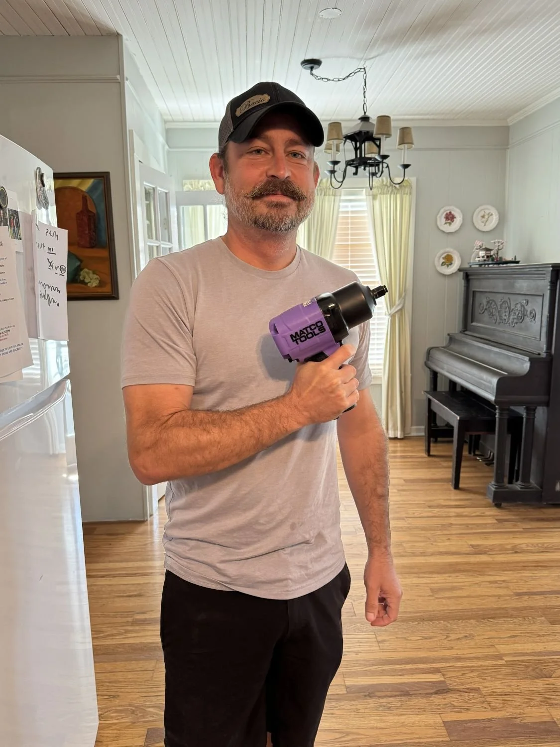 Man with a mustache and beard holding a purple Matco Tools pneumatic impact wrench in a home kitchen, wearing a light gray t-shirt and a black cap.