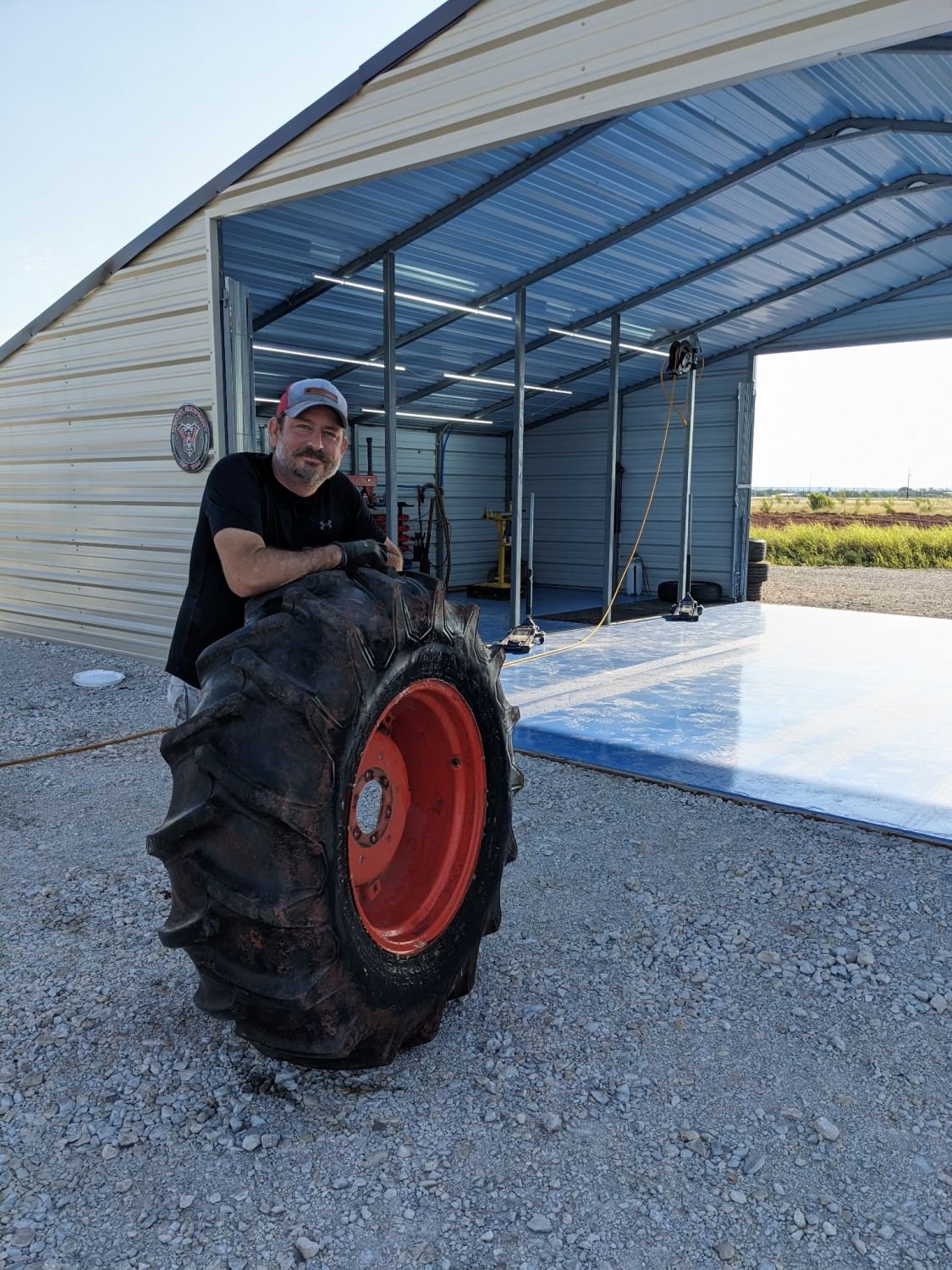 Man with beard and cap standing outside of a metal building with a large tractor tire in front of him, on a gravel surface, with open rural landscape in the background.