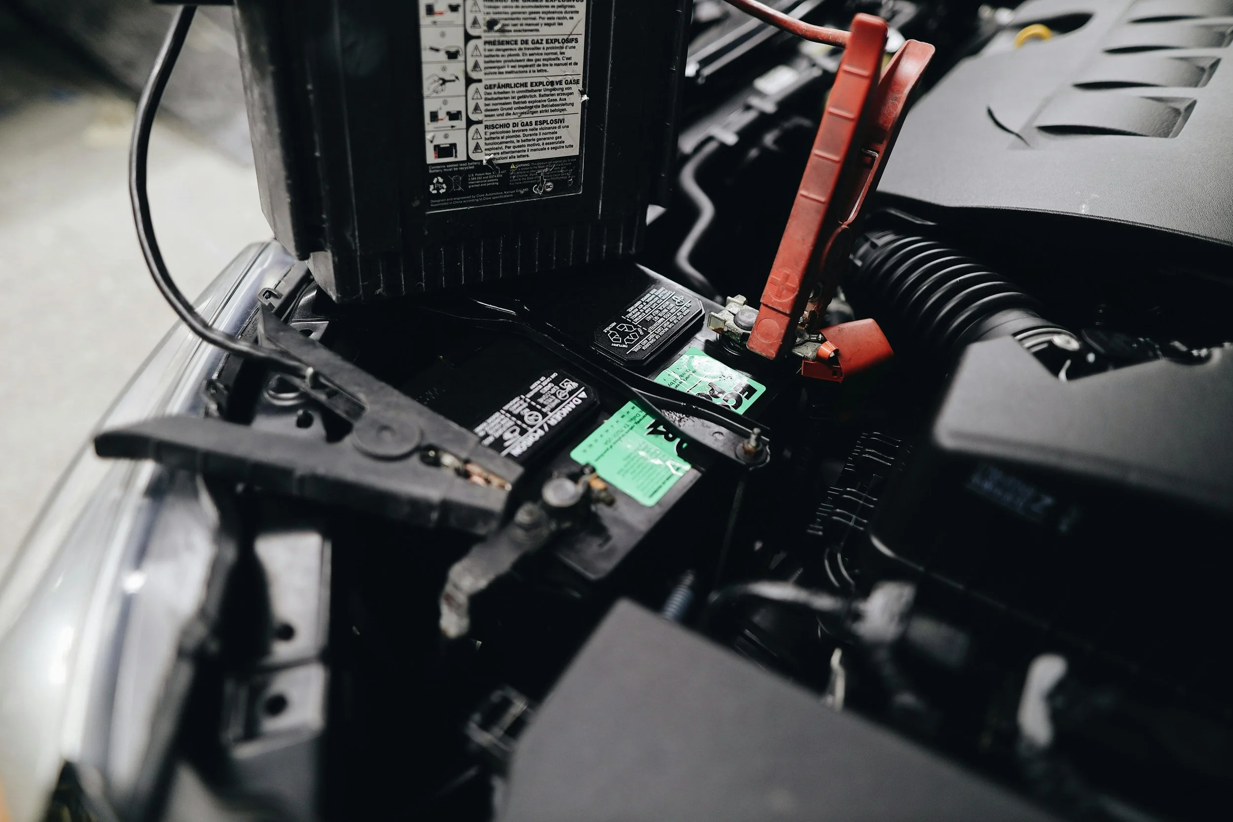 Close-up of a car battery with warning labels and a red terminal connector, inside a vehicle engine compartment.