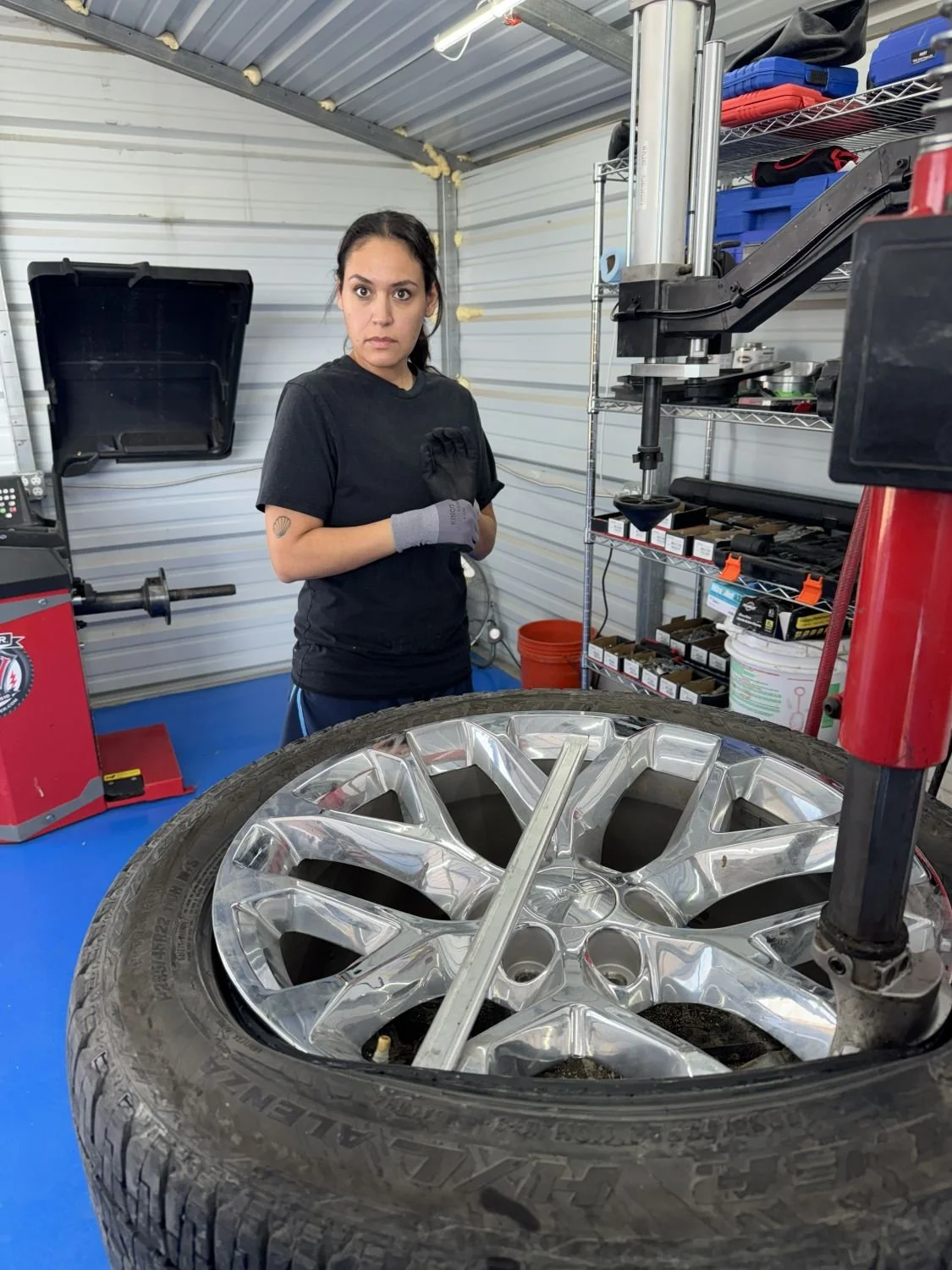 A woman in black shirt and gloves stands in a garage next to a tire mounted with a shiny alloy wheel, with tools and shelves in the background.