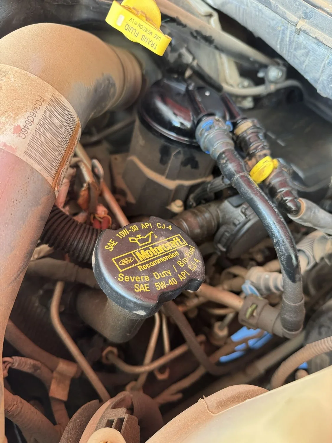 Close-up of a vehicle engine showing oil cap, hoses, and various engine components, with a yellow label on a part in the top area.