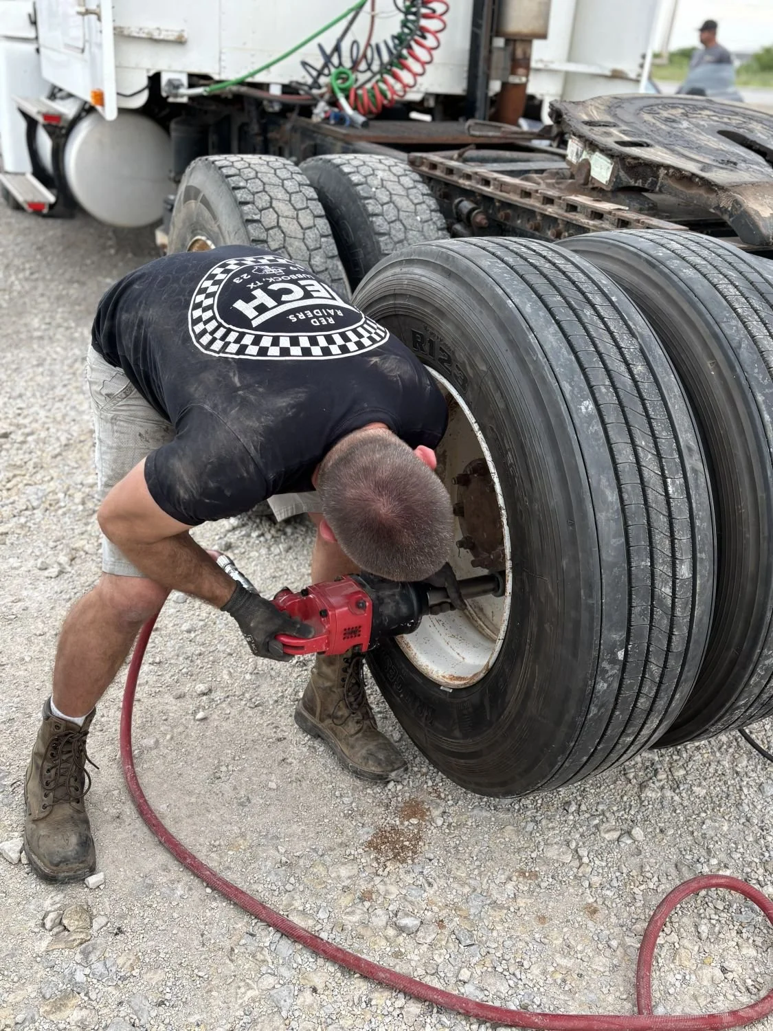 A man working on a truck tire using a power tool outdoors, with several tires stacked nearby, and a truck cab visible in the background.
