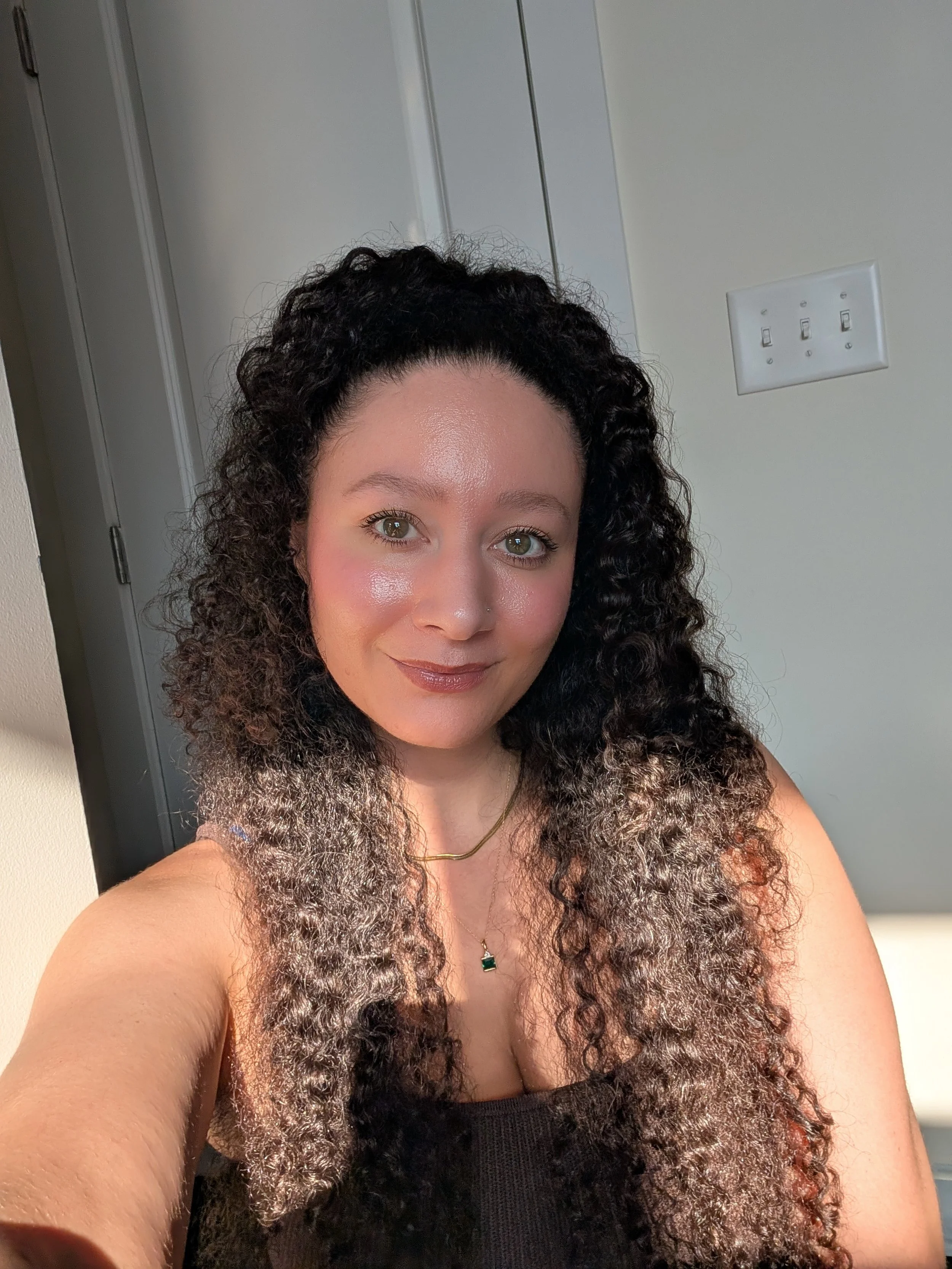 A woman with curly hair and a glowing complexion taking a selfie indoors, with a light switch and a plain wall visible behind her.