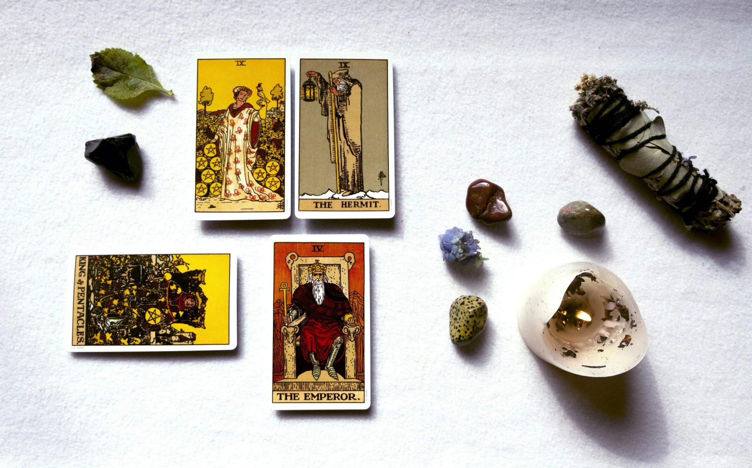 Three tarot cards laid out on a white surface, with various stones, leaves, and a smudging stick nearby. The visible cards are the Nine of Pentacles, The Hermit, and The Emperor.