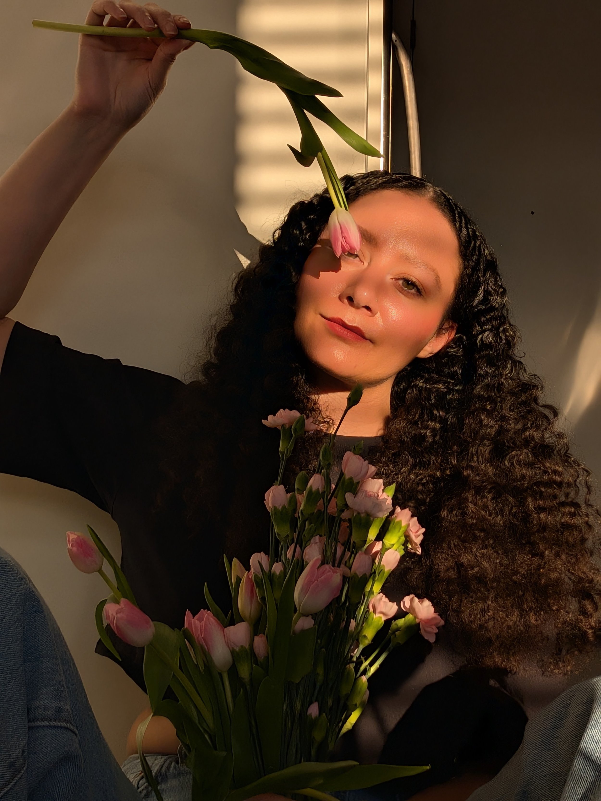 A woman with dark, curly hair holds a bouquet of pink tulips and underwater plants. She is partially submerged in water, with sunlight casting reflections on her face and the background.