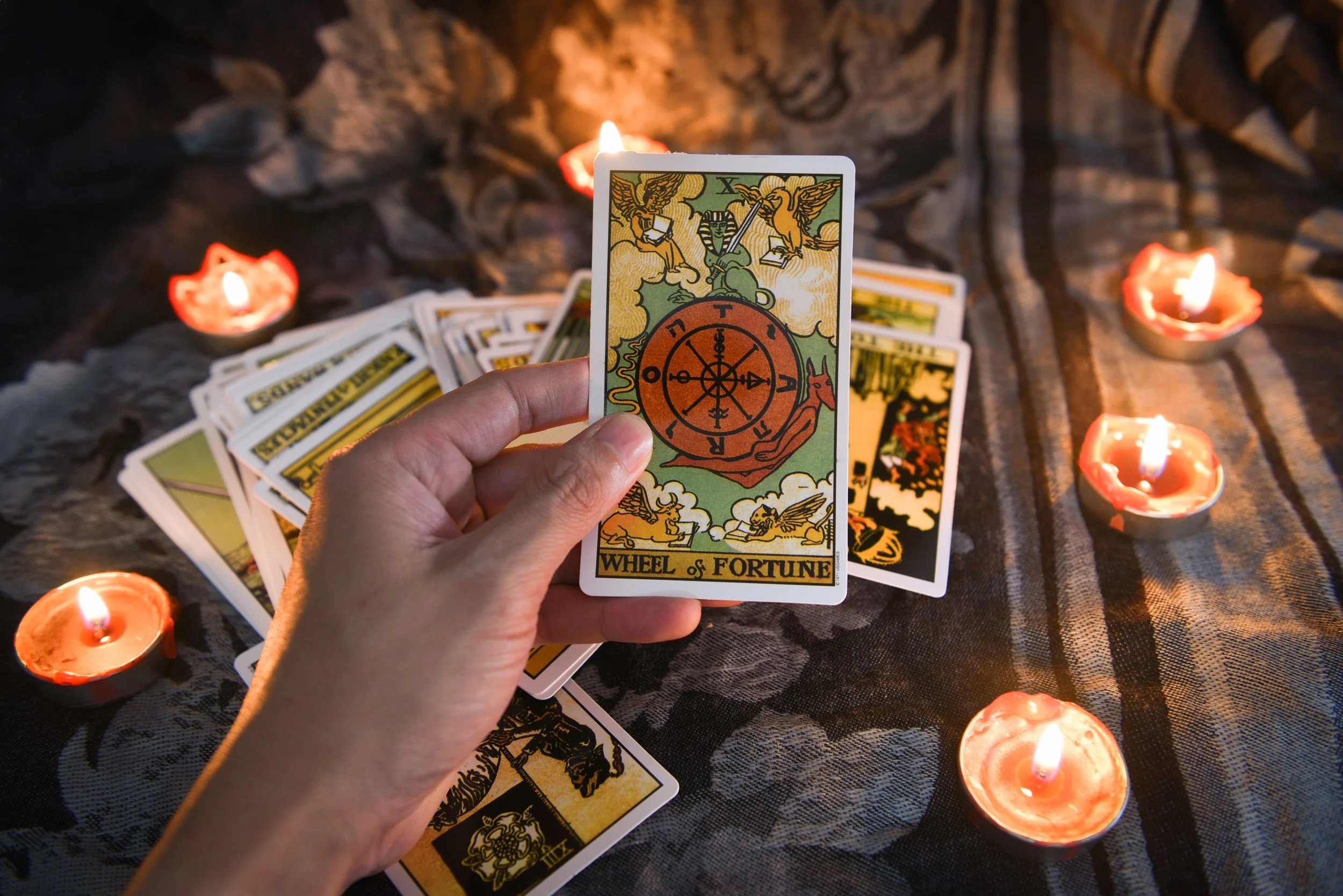 A person holding a tarot card, 'Wheel of Fortune', above scattered tarot cards on a dark cloth, with small lit candles around.