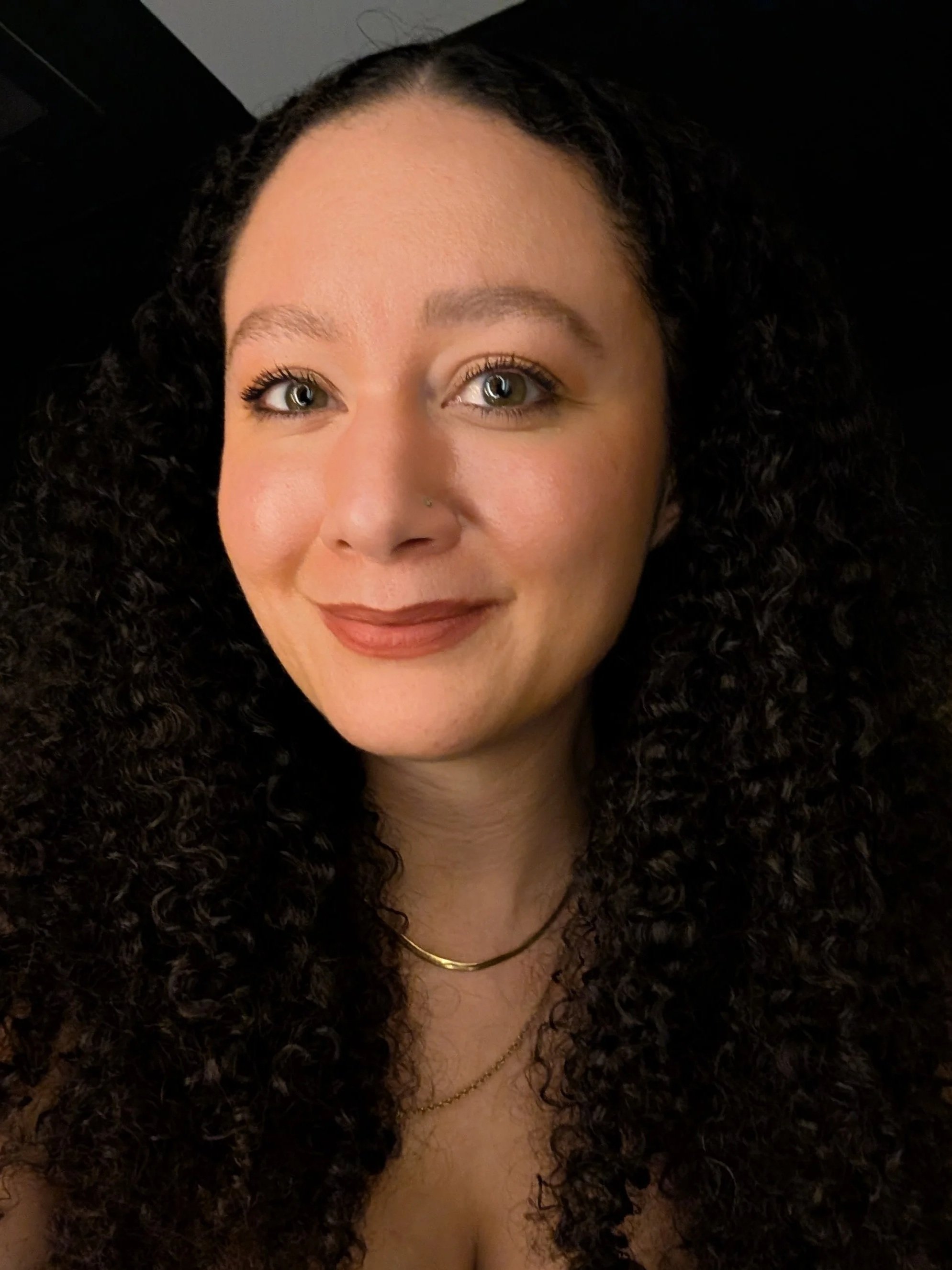 Close-up of a woman with dark curly hair, wearing makeup, gold necklaces, and a small nose piercing, smiling slightly.