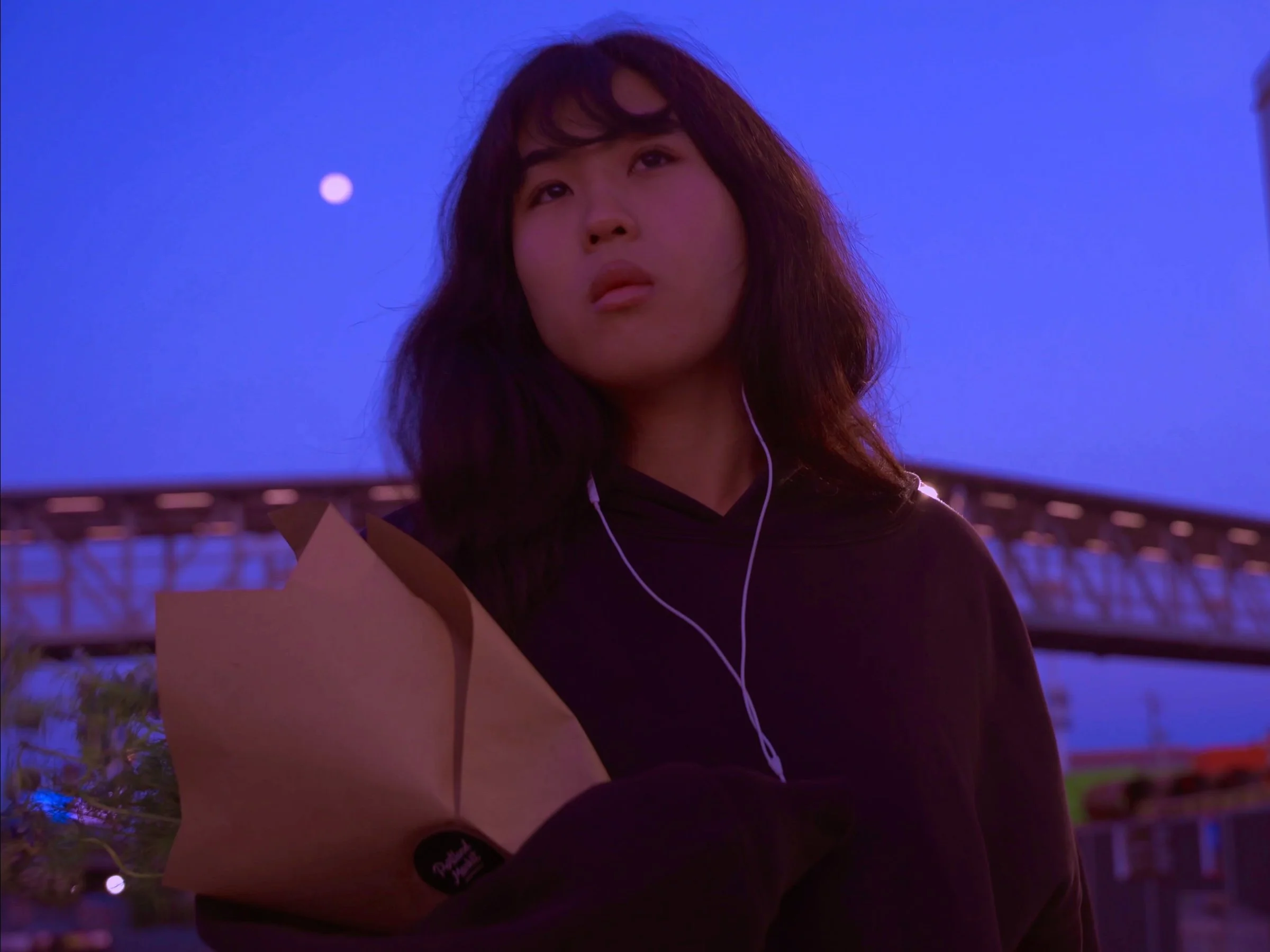 A young woman with long dark curly hair and a serious expression, wearing a black hoodie, white earbuds, and holding a flower bouquet, stands outdoors under a vibrant blue sky at dusk.