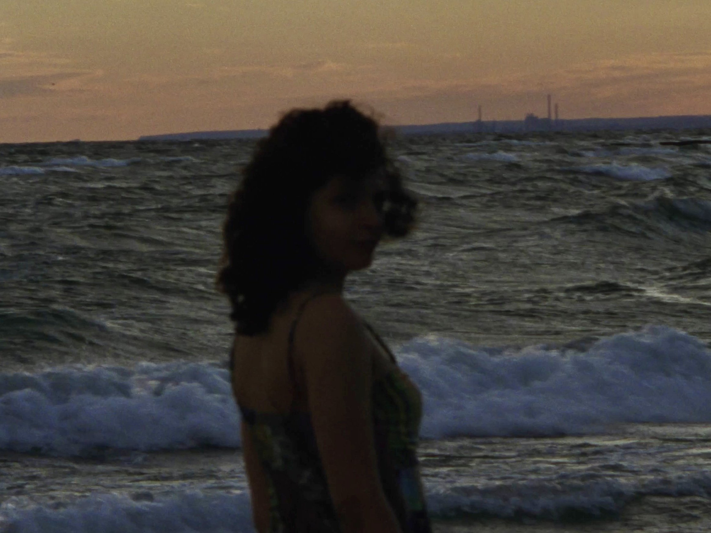 A woman with curly hair standing on a beach at sunset or dusk, with waves and an industrial harbor in the background.