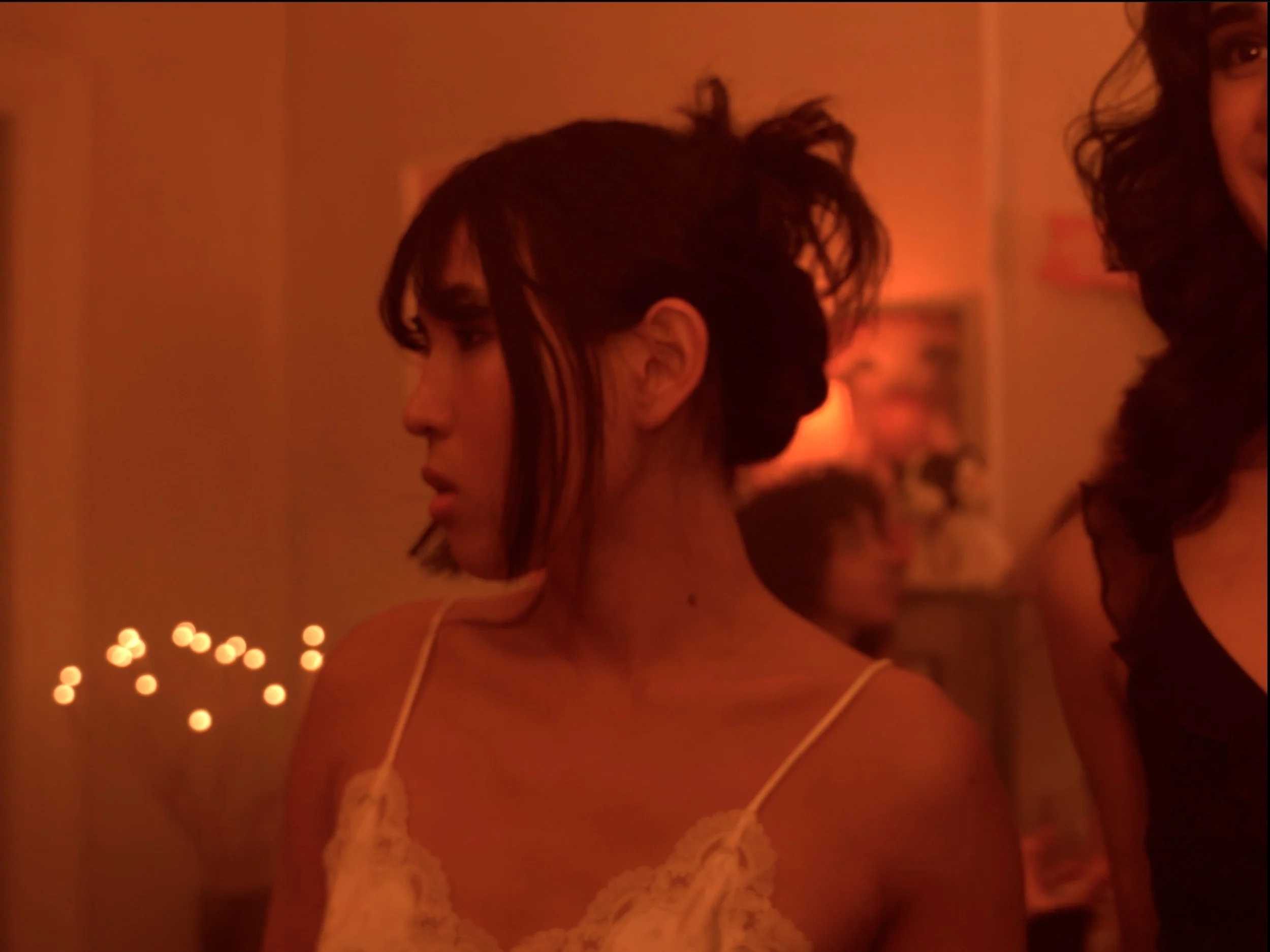 Close-up of two women with dark hair, one with a high messy hairstyle, wearing lingerie, in a dimly lit room with red lighting and string lights in the background.
