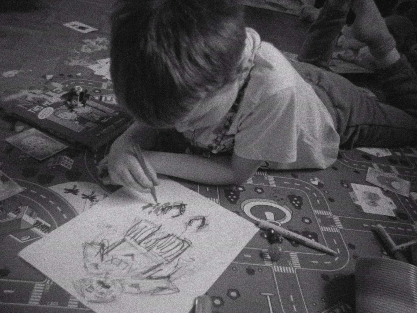 A young boy lying on a colorful play mat, drawing with crayons on a piece of paper, surrounded by toys and books.