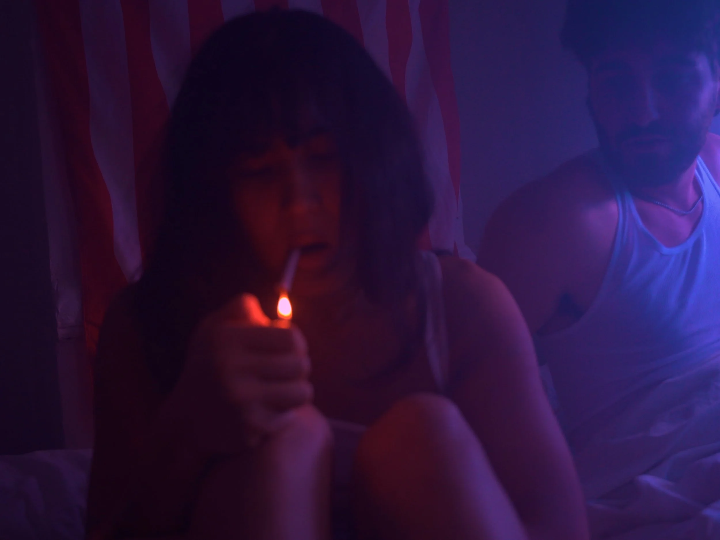 A young woman and young man in a dimly lit room with purple and blue lighting. The woman is smoking a cigarette, and the man appears to be staring at the woman.
