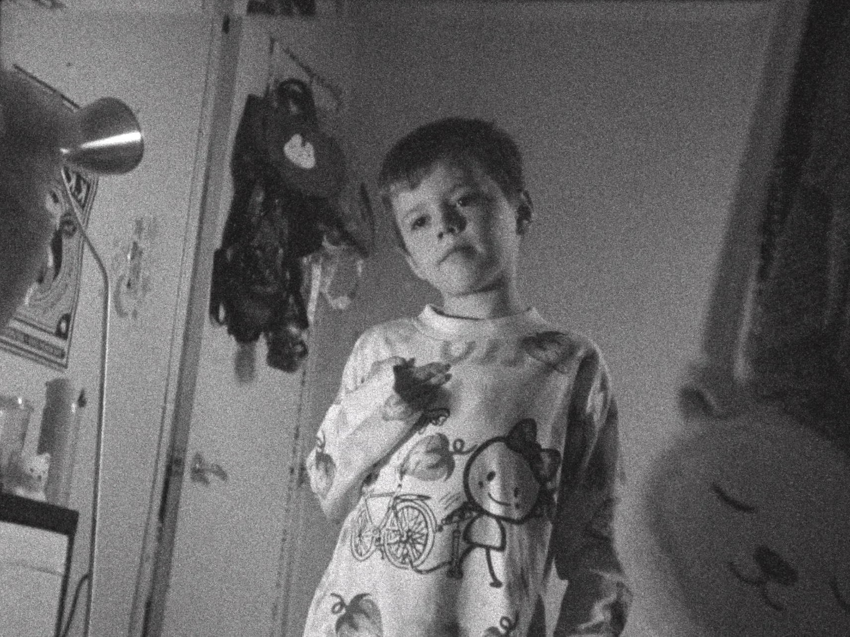 A young boy with short hair standing in a room, wearing a longsleeve with cartoon characters on it, looking at the camera with a neutral expression.