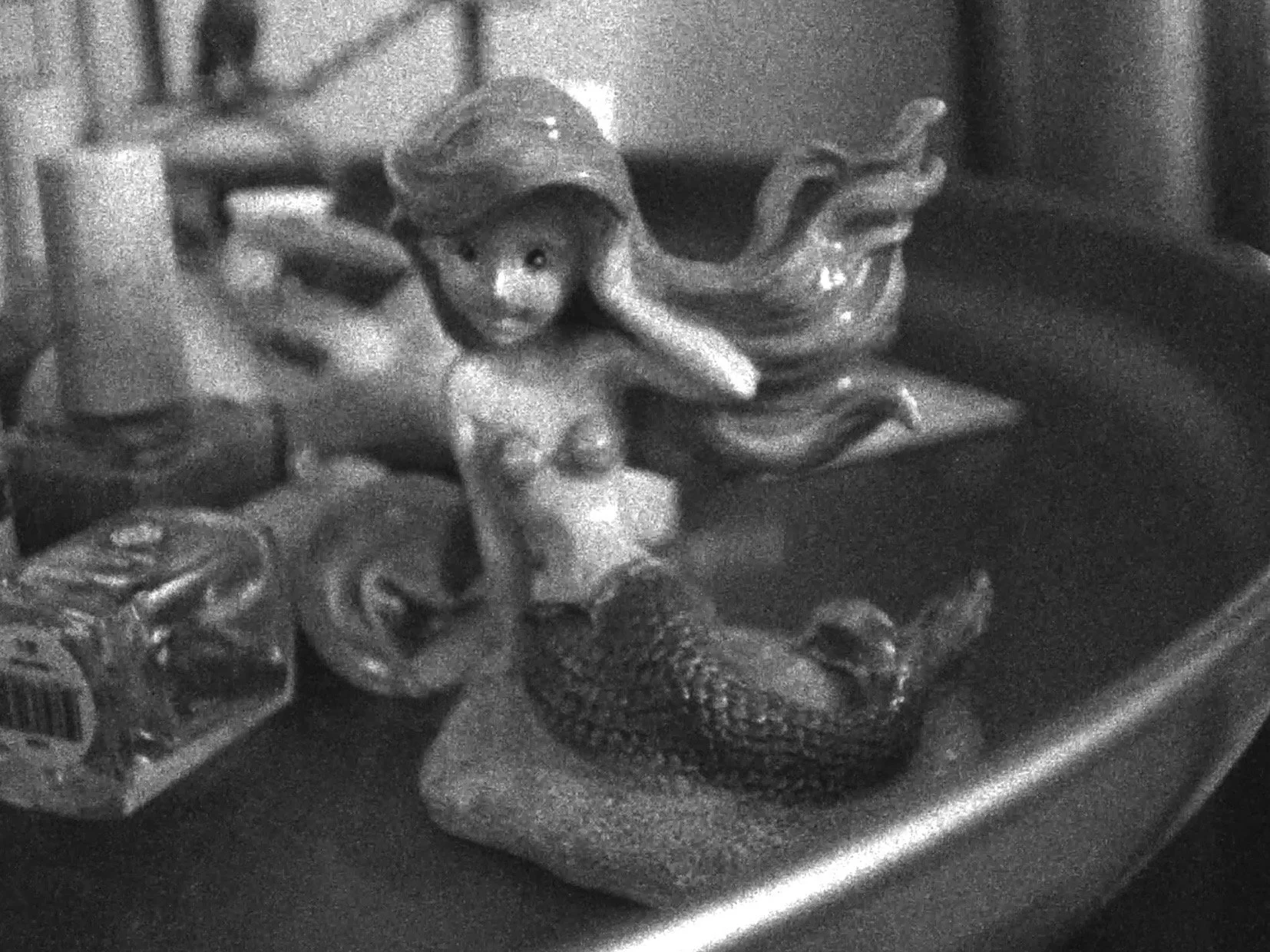 A ceramic mermaid figurine, next to a ceramic octopus that appears, placed on a table with other objects in the background, like nail polish.
